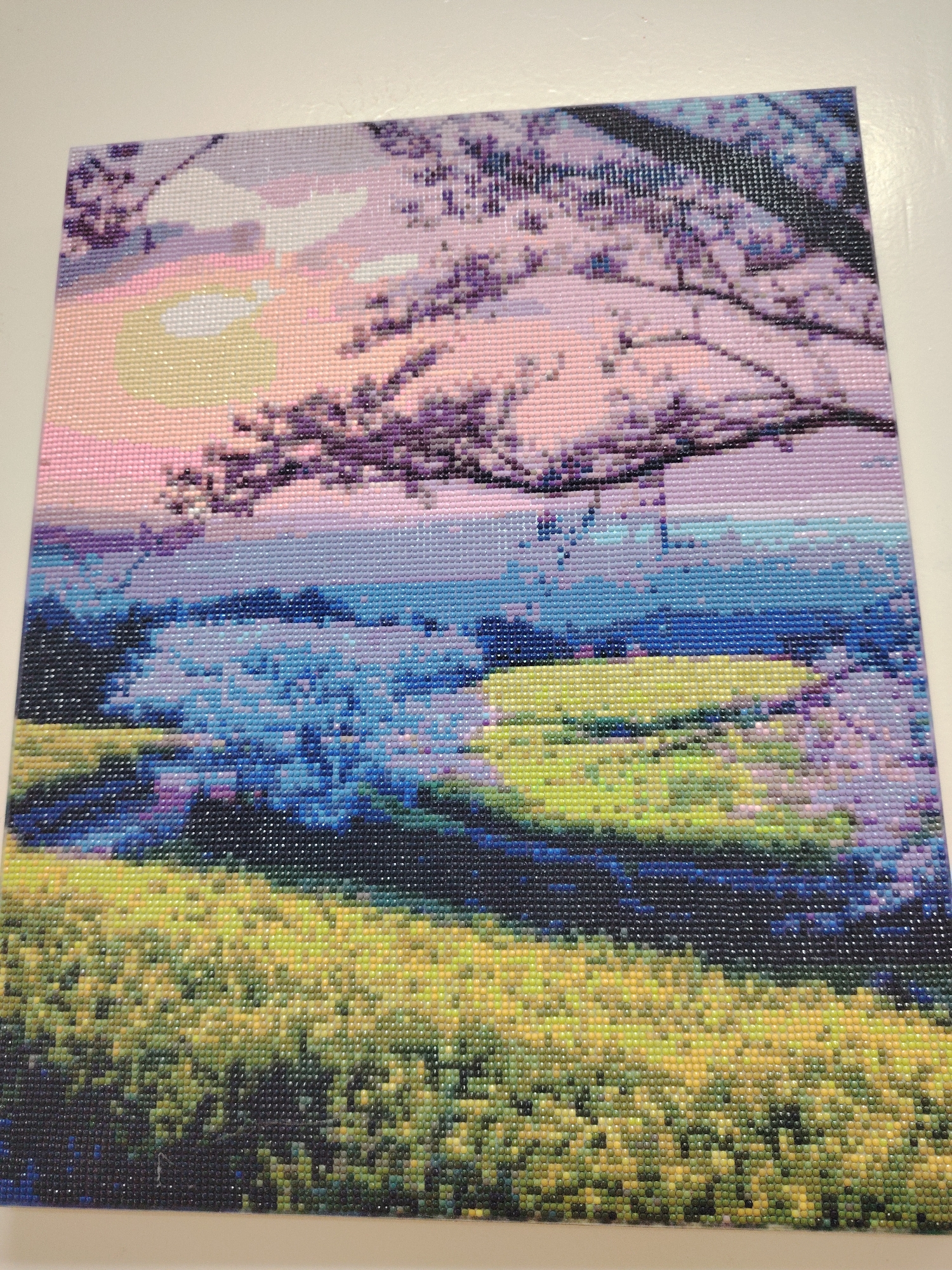 The image is a square diamond painting depicting a landscape. The top portion shows a pink and white sky with a yellow sun, partially obscured by tree branches with purple blossoms. Below the sky, there's a body of water rendered in shades of blue, followed by a green field that transitions to yellow in the foreground. The painting is composed of small, square, shiny tiles. The scene does not depict the city of Sarapul, Russia.