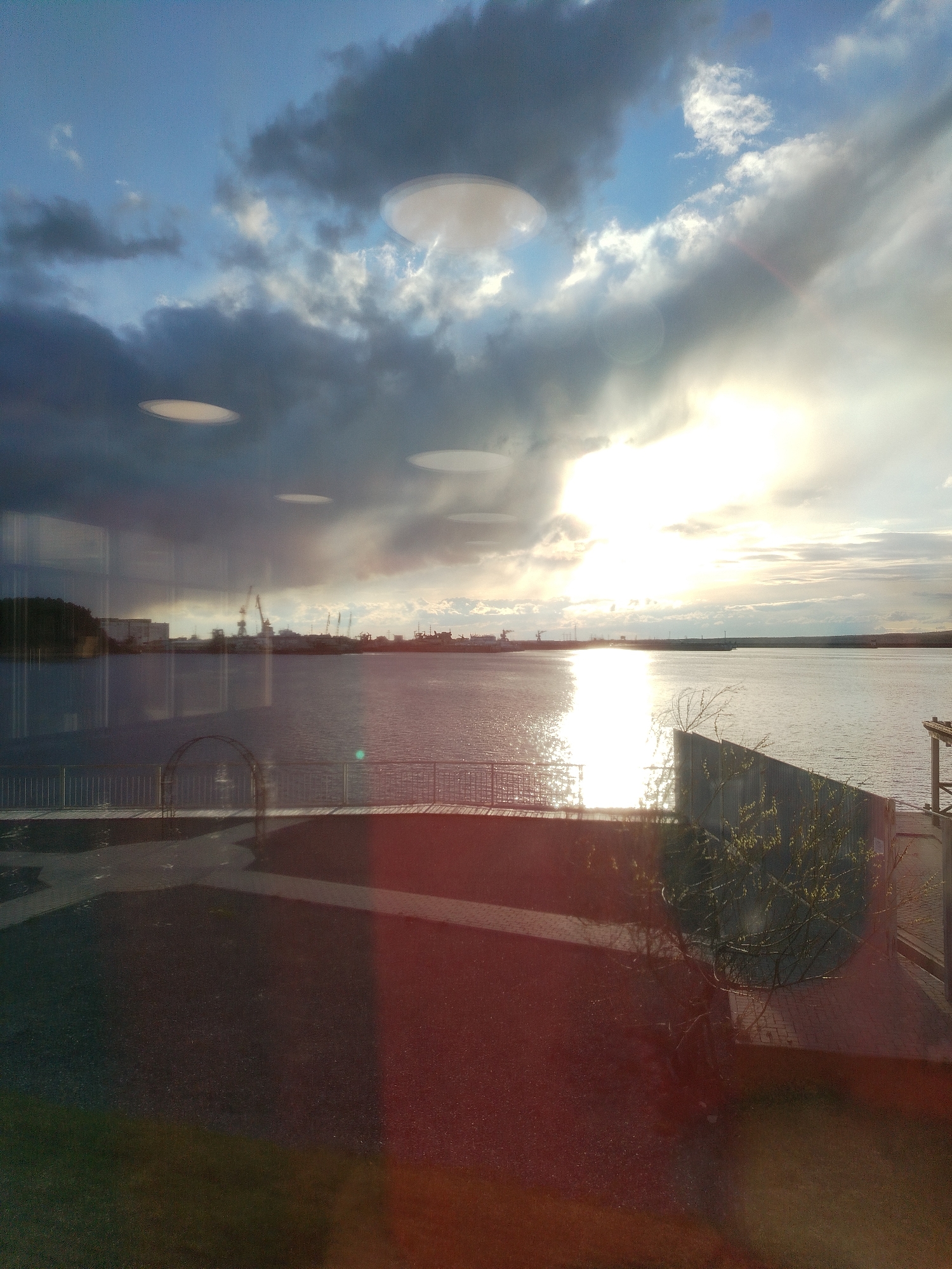 The image captures a dramatic sunset or sunrise over a wide body of water, likely the Kama River in Chaikovsky, Russia. The sky is dominated by large, dark, and heavy clouds, particularly on the left side, with patches of bright blue and white clouds visible where the sun breaks through. The low sun on the right side of the frame casts a brilliant, intense light that creates a shimmering, almost blinding reflection across the surface of the water.
The scene appears to be viewed through a window, evidenced by several circular reflections of interior lights visible in the sky and mid-ground. Reflections of the immediate foreground, including a path, a low metal fence, and a small decorative archway, are superimposed over the outdoor scene.
On the distant shore across the expansive water, industrial elements are visible, including numerous cranes and several buildings, suggesting a port or a commercial waterfront. A dark, hilly landmass is also present on the far left bank. The foreground shows a paved area with some sparse, bare vegetation near the fence line. The bottom and right sections of the image are tinged with a reddish-orange hue, possibly due to lens flare or reflections on the window glass.
No people are visible, and no specific activities are taking place other than the natural light display. The image effectively blends the interior perspective with the grand outdoor landscape, characterized by dynamic weather and striking natural light. No visible text is present.