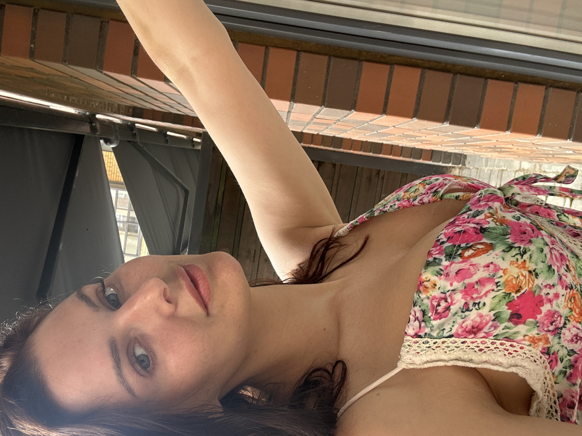 A woman with brown hair, fair skin, and light-colored eyes is pictured in a close-up, upper-body shot. She is wearing a floral print dress with pink and white flowers. Her left arm is extended outwards, and her right shoulder is visible. The background consists of a brick wall and wooden siding. The image appears to be taken outdoors during the day.