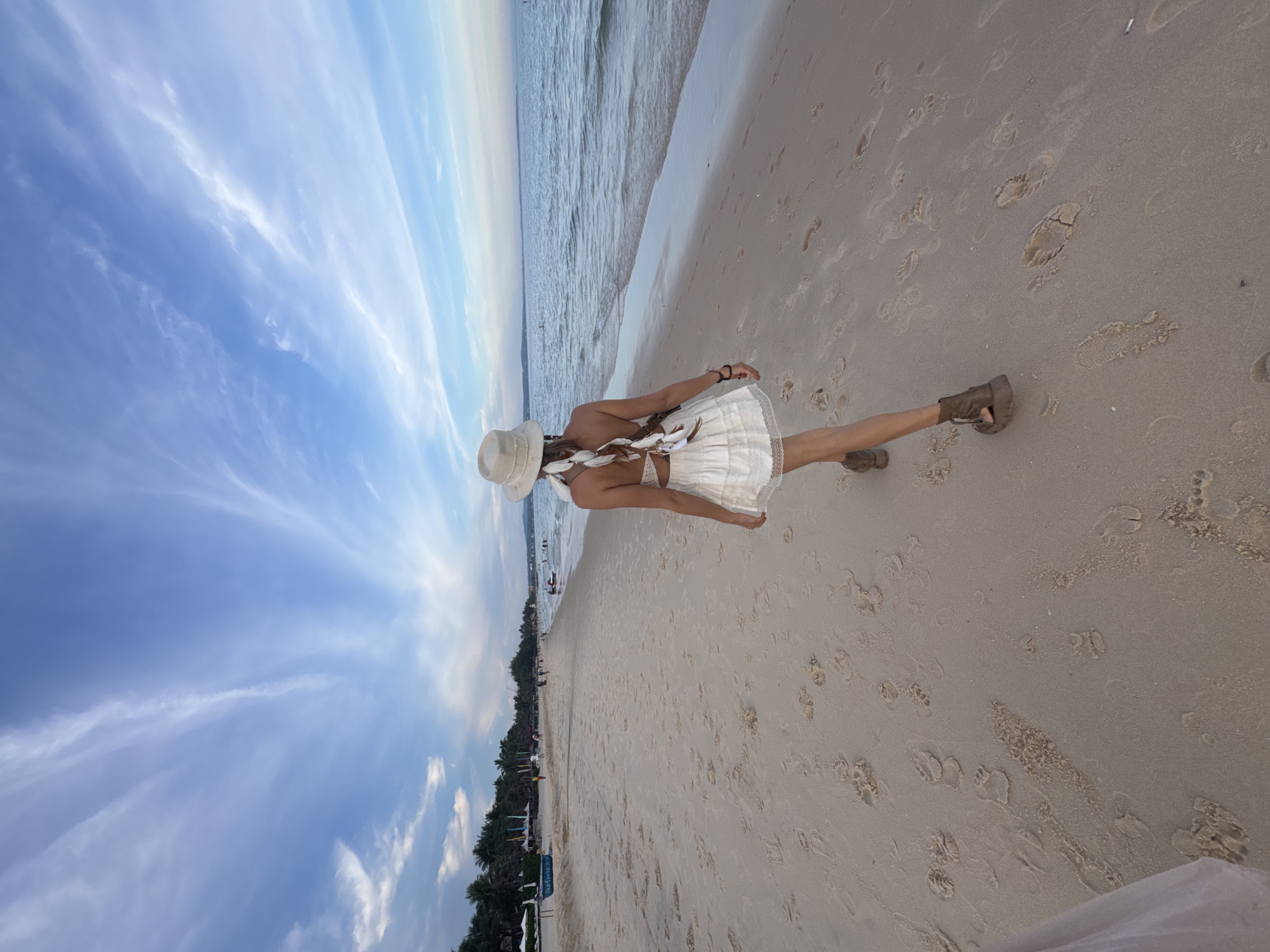 A woman in a white hat and a white, ruffled, short-sleeved dress walks on a sandy beach in Phu Thuy Ward, Vietnam. Her back is to the camera, and her hair is styled in a braid decorated with white feathers. She wears brown, open-toed boots. Her footprints are visible in the sand, along with many other footprints. To her left, the sea stretches out to the horizon, and the sky is blue with wispy white clouds. Further down the beach to the left, there are palm trees, lounge chairs, and a few people. The overall scene is serene and tranquil.