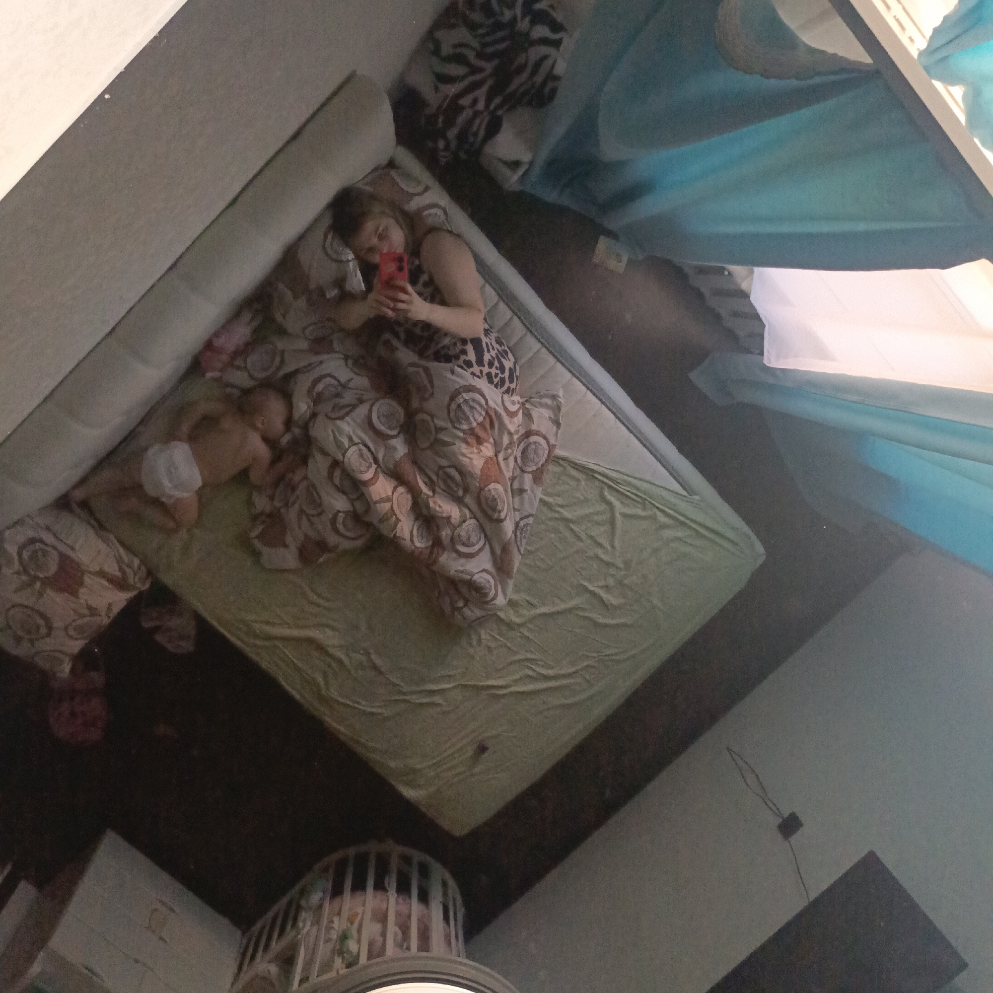 The image displays an indoor scene, likely a reflection, captured from an elevated and tilted perspective. A woman with light brown hair is lying on a mattress placed directly on a dark floor. She is wearing a patterned sleeveless top and is holding a red smartphone in front of her, appearing to be taking a selfie or looking at the screen. Beside her, a baby wearing a white diaper is sleeping on its stomach. Both are partially covered by a patterned blanket featuring circular, possibly coconut-themed, designs. A light green fitted sheet covers another part of the mattress.
In the upper right section of the frame, light blue fabric or curtains are draped. The bottom central area shows a portion of a white slatted crib or playpen, with some colorful elements visible inside. The room features a dark floor and a light-colored wall or baseboard along the left edge. A small, dark, square object with a thin wire is attached to a wall in the lower right. The scene conveys a calm, domestic atmosphere, likely during a relaxed moment. There is no visible text in the image.