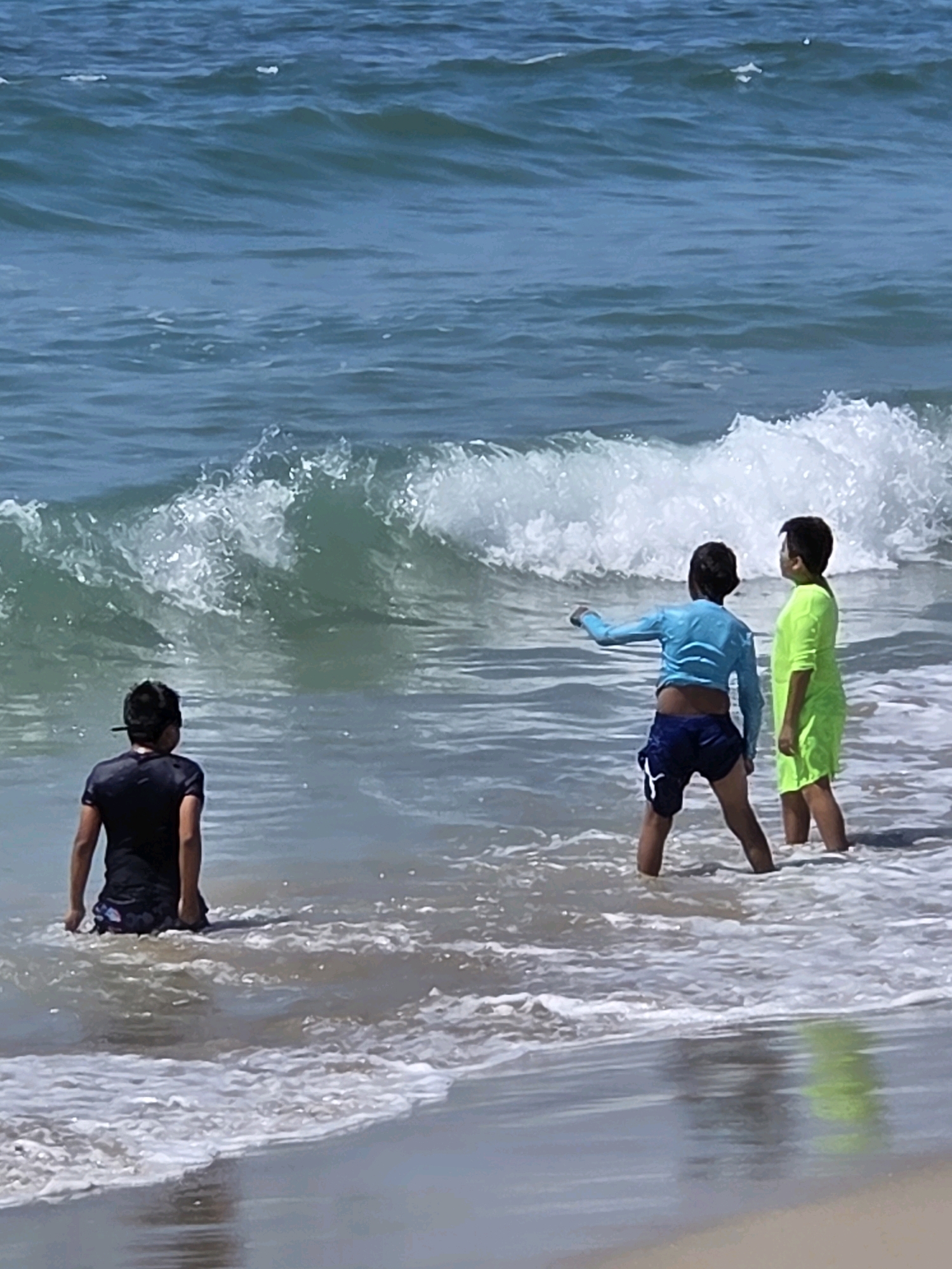 Three young boys are in the shallow water at the beach, facing a breaking wave. On the left, a boy in a black t-shirt and patterned swim trunks stands with his back to the camera. In the center, a boy in a blue long-sleeved rash guard and navy blue swim trunks points towards the wave. To the right, a boy in a neon green short-sleeved shirt and swim trunks stands looking at the wave. The ocean water is turquoise and has white foam from the breaking wave. The scene is set on a sandy beach, under a clear blue sky. The location is the beach in Santa Monica, California, United States of America.