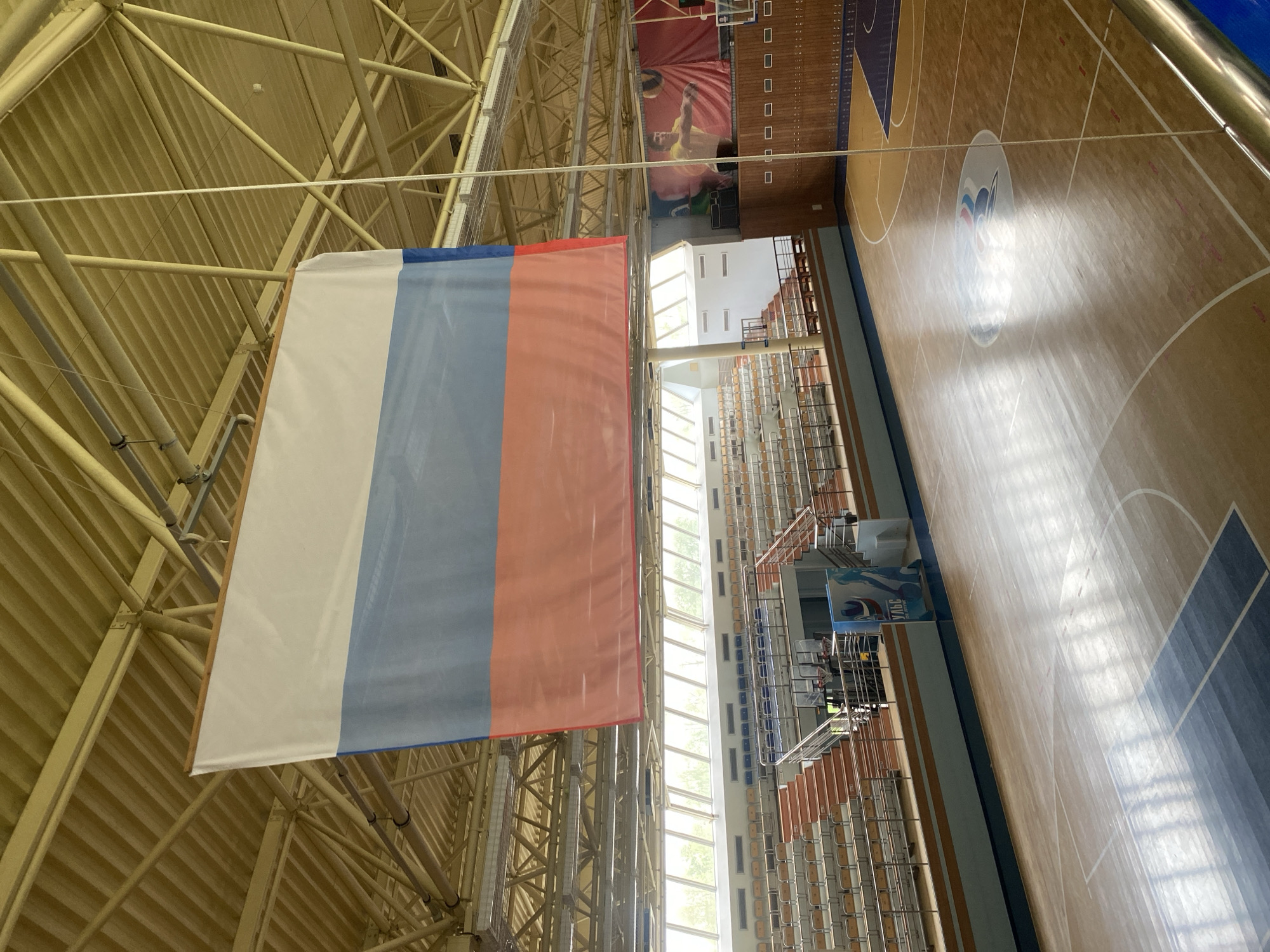 The image shows the interior of the Sports Complex "Sayan", located in Chaikovsky, Russia. A large flag of Russia hangs from the ceiling. The space includes a wooden basketball court with painted lines and the logo of the sports complex in the center. Spectator seating is visible. The building's architectural features include exposed metal beams and supports in the ceiling, and a tall bank of windows running along the side of the court. A banner depicting athletes playing volleyball hangs above the seating.