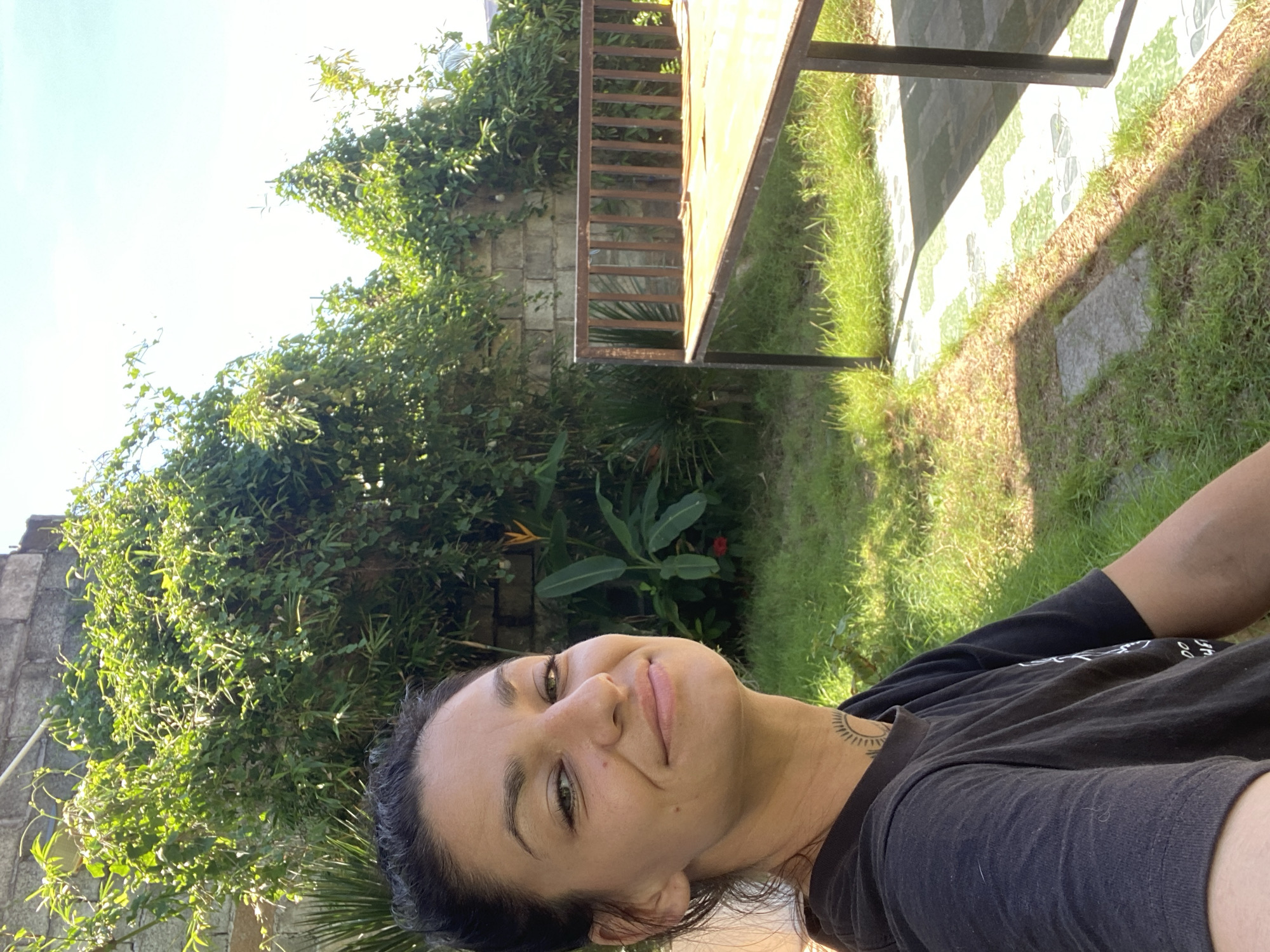 A woman is taking a selfie in an outdoor setting. She has dark hair pulled back and is wearing a black t-shirt with a tattoo on her neck. She is smiling at the camera. The background consists of a brick wall covered in lush green vegetation. To the right, there is a table with a wooden top and metal legs, positioned on a paved area with grass. The overall scene suggests a sunny day in a garden or courtyard. The location is identified as Mui Ne, Vietnam.