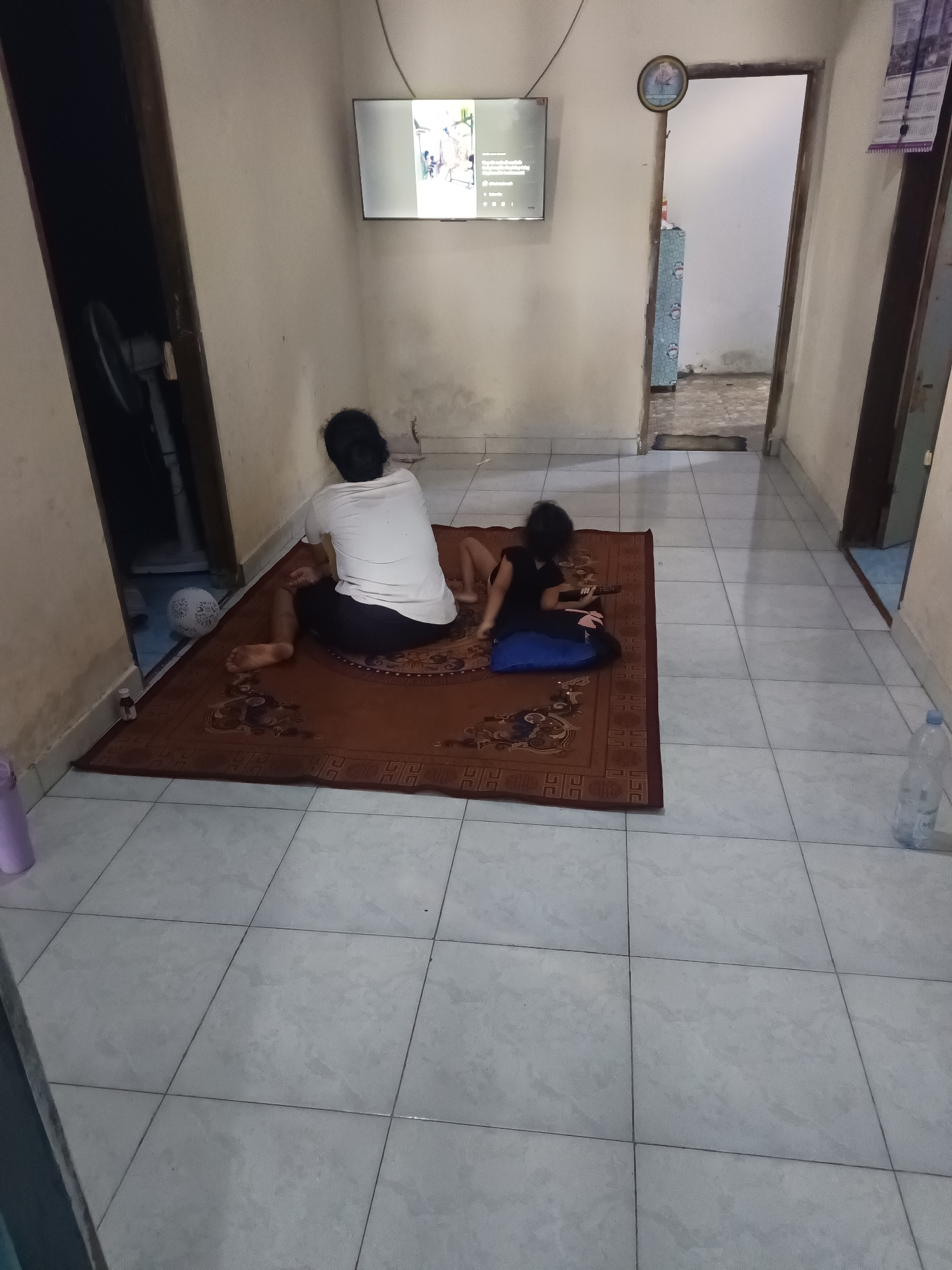 The image depicts an indoor scene, likely a home, in Terbanggi Marga, Indonesia. Two individuals are present on a brown rug placed on a tiled floor. The person on the left is an adult, seated with their back to the camera, wearing a white t-shirt and dark pants. Their legs are crossed and extended. To the right, a child is also seated, with their back to the camera, wearing a black top and pink leggings. The child is leaning on a blue cushion and appears to be holding a small, dark object, possibly a toy or an electronic device.
The room has beige walls and a light-colored tiled floor. A flat-screen television is mounted on the wall to the left, displaying an image. To the far left, partially visible through a doorway, is a dark area with what appears to be a standing fan and a white ball with a circular pattern. On the right side of the image, another doorway leads to an adjacent room. A water bottle is visible on the floor to the right of the rug. A clock is mounted on the wall above the doorway on the right. The overall environment appears to be a modestly furnished living space.