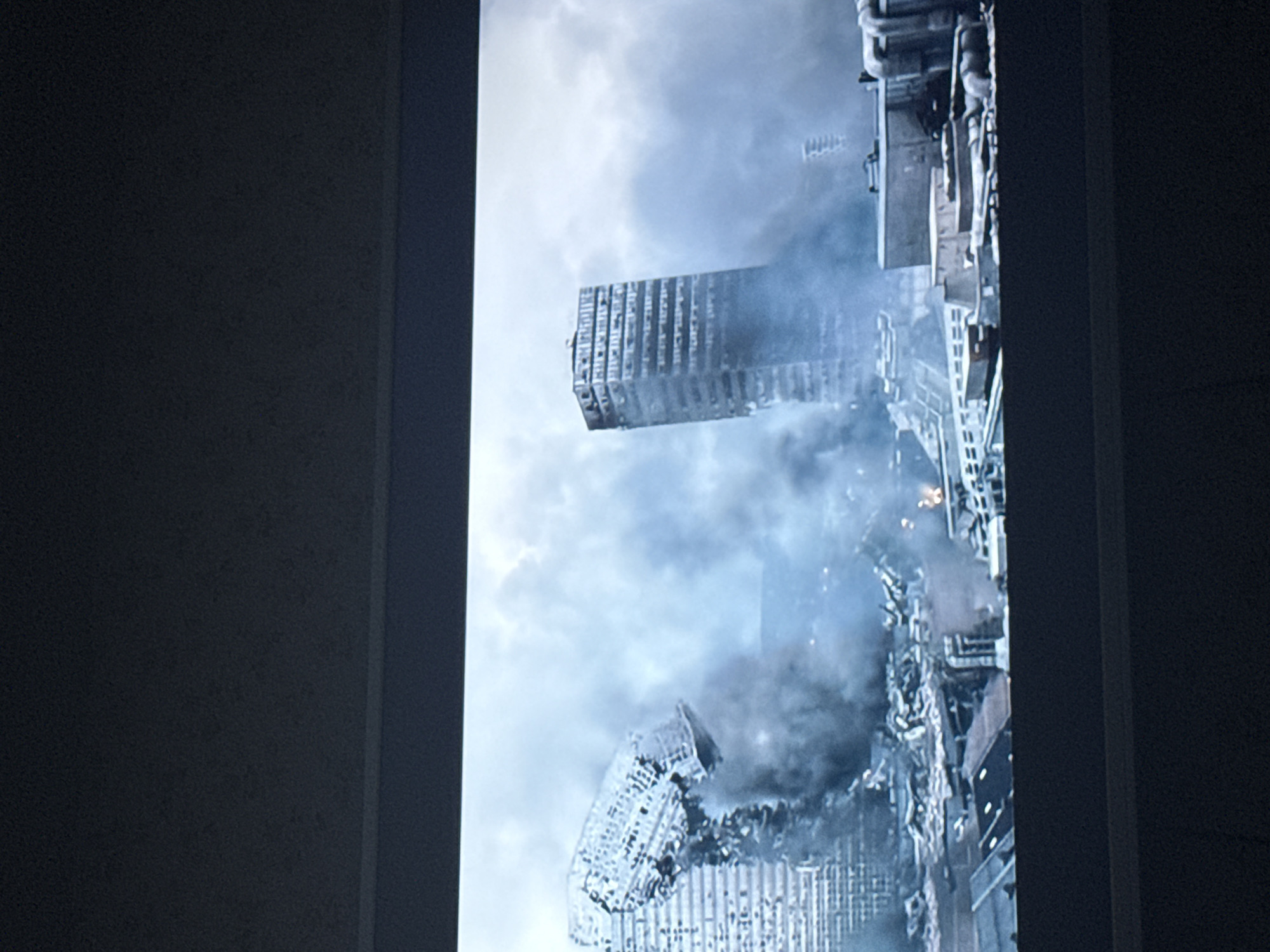 The image shows a cityscape viewed through a rectangular opening, possibly a window. The city is in a state of destruction, with damaged buildings and smoke or dust obscuring the view. The buildings are tall and appear to be of a modern design. There are no people visible in the image. The environment is bleak and filled with debris. The location cannot be confirmed as Mytishchi, Russia based on the provided image.