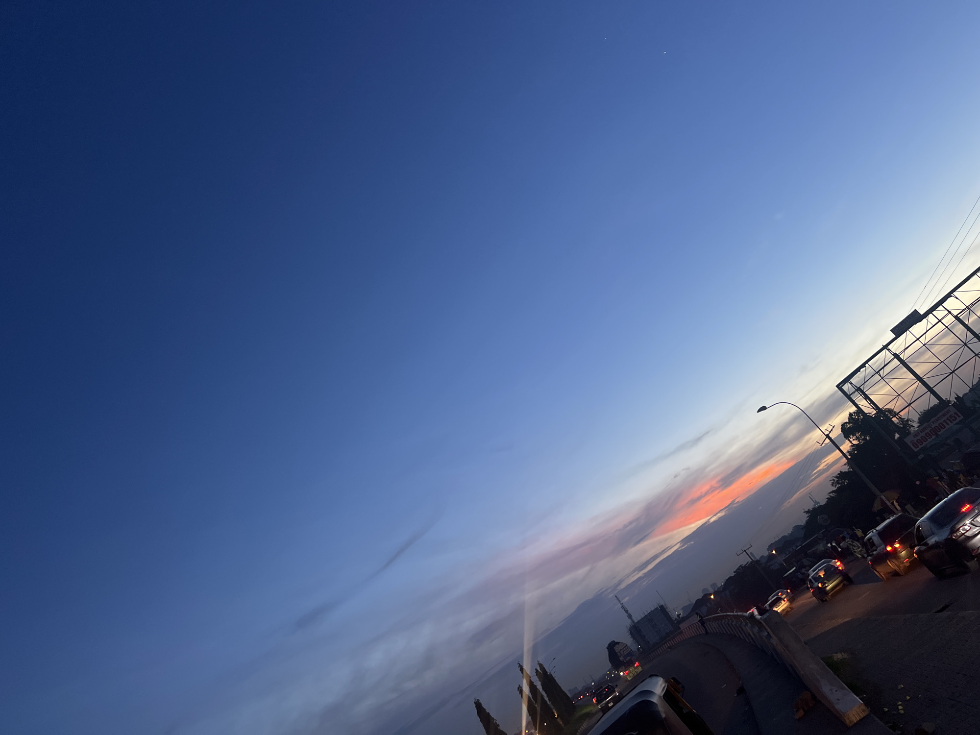 The image displays an urban twilight scene, likely at sunset, over a roadway in Aso, Nigeria. The sky dominates the upper two-thirds of the frame, exhibiting a color gradient from a deep indigo blue in the upper left quadrant, transitioning to a lighter blue, and then to a band of orange and pink hues along the horizon on the right. A faint crescent moon is visible in the upper left, and minimal cloud cover is present.
The lower third of the image shows a multi-lane road, possibly an elevated overpass or bridge, curving from the lower left towards the mid-right. Multiple vehicles, primarily passenger cars, are on the roadway with their headlights and taillights illuminated, indicative of low light conditions and active traffic.
On the right side of the frame, a large metal-framed billboard structure stands silhouetted against the sky. Text partially visible on the billboard includes "MARKETI..." and "ABU..." along with a contact number, "09090001151." An illuminated streetlight extends over the roadway from the right side. In the background, various buildings, some with visible construction elements, are discernible. Trees and other foliage line the roadside and appear in the distance. Concrete barriers delineate parts of the road. Scattered light sources from buildings and vehicles illuminate the scene, consistent with an urban environment in a Nigerian city such as Abuja (where Aso is located) during dusk.