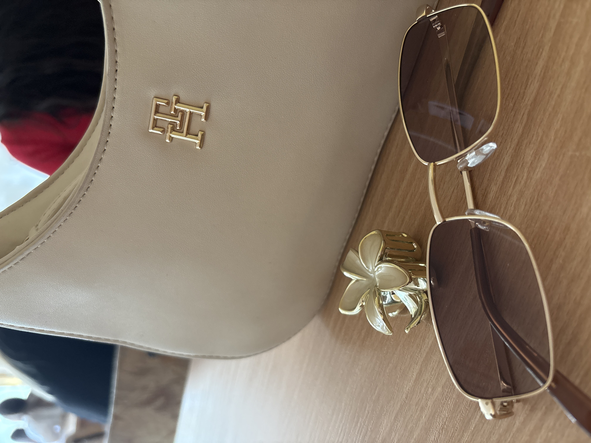 The image features a beige Tommy Hilfiger purse with a gold logo, a gold flower hair clip, and a pair of brown-tinted sunglasses with gold frames, all situated on a wooden surface. The location is not discernible from the image. There are no people visible in the frame, although it appears someone is partially in the background on the lower left of the image. The subjects are the purse, hair clip, and sunglasses. There are no visible actions or interactions. The environment is a simple wooden surface.