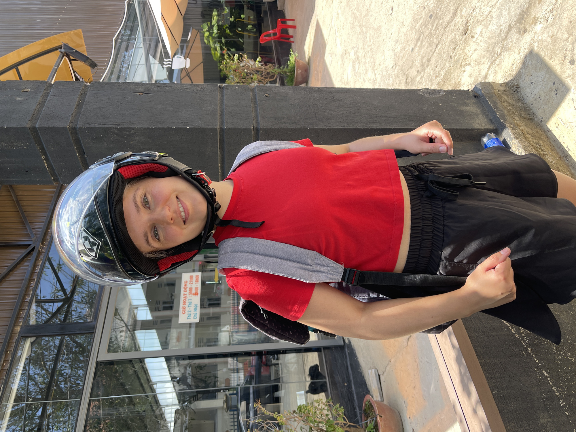 A young woman wearing a full-face motorcycle helmet, a red crop top, and black shorts stands outside a building with a glass facade in Da Lat, Vietnam. She is also wearing a gray backpack. The building's exterior features black beams and a sign displaying business hours in Vietnamese. A potted plant sits near the building's base. Sunlight is visible, casting shadows on the concrete ground. A water bottle is on the ground near her.
