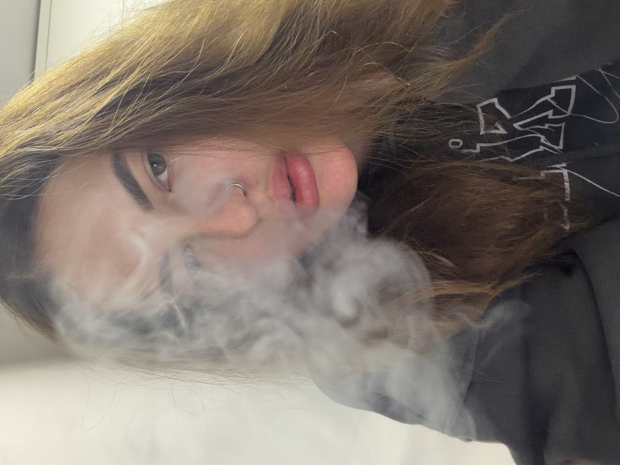 A close-up portrait shows a young person with long, light brown hair, possibly with blonde highlights, tilted to the right in the frame. A dense cloud of white smoke or vapor billows from their mouth area, partially obscuring the lower face and drifting towards the foreground.
The visible eye is green, complemented by well-defined, dark eyebrows. A silver hoop nose ring adorns the left nostril. The lips are full and reddish, slightly parted. The person is wearing a dark-colored garment, likely a hoodie or sweatshirt, on which a stylized white graphic composed of angular shapes, possibly abstract letters, is visible.
The setting appears to be indoors with artificial lighting and a plain, light-colored background. The person's expression is neutral and direct, gazing towards the viewer through the smoke. The image captures the act of exhaling smoke or vapor, suggesting the use of a vape or similar device. The original image is rotated 90 degrees clockwise. There are no specific visual cues to indicate the time of day, weather, or the location of Krasnodar, Russia.