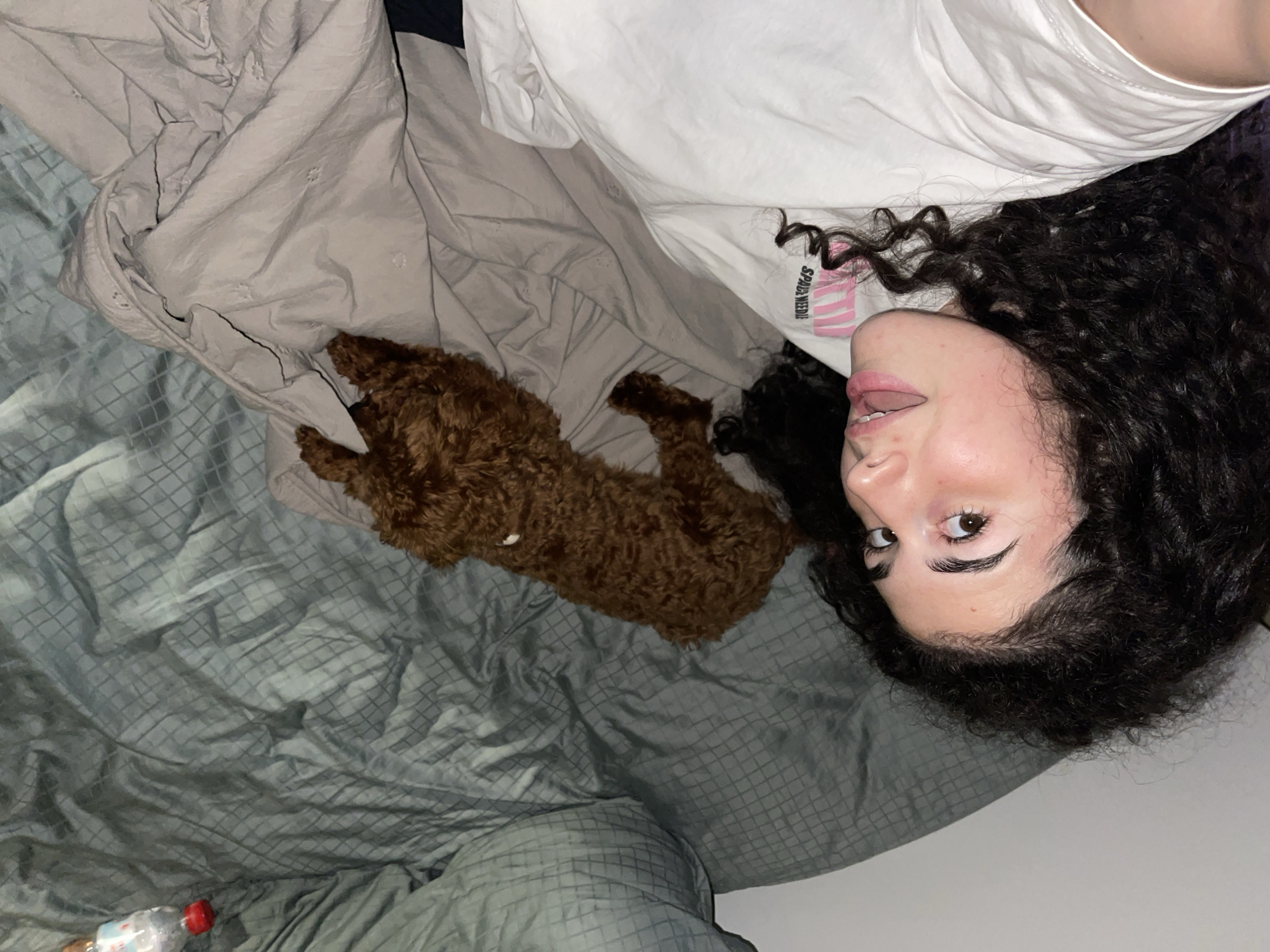 The image captures a young woman and a small, brown, curly-haired dog relaxing on a bed in an indoor setting, contextually located in Sofia, Bulgaria. The perspective is from above, with the image rotated, showing the woman's head on the right side of the frame, looking directly at the camera with a soft, relaxed expression. Her dark, curly hair frames her face, and she has visible winged eyeliner and a subtle pink lip tint, along with a ring on her finger.
She is wearing a white t-shirt, which has the black text "SPACE NEEDLE" printed on it, accompanied by pink graphic elements. To her left, nestled on a light grey blanket, is the dog, curled up and appearing to rest. The bed is covered with a mix of light grey, possibly quilted, blankets and a dark grey or bluish-grey patterned sheet. In the bottom-left corner of the frame, a clear plastic water bottle with a red cap is visible on the patterned sheet. The overall ambiance is quiet and relaxed.