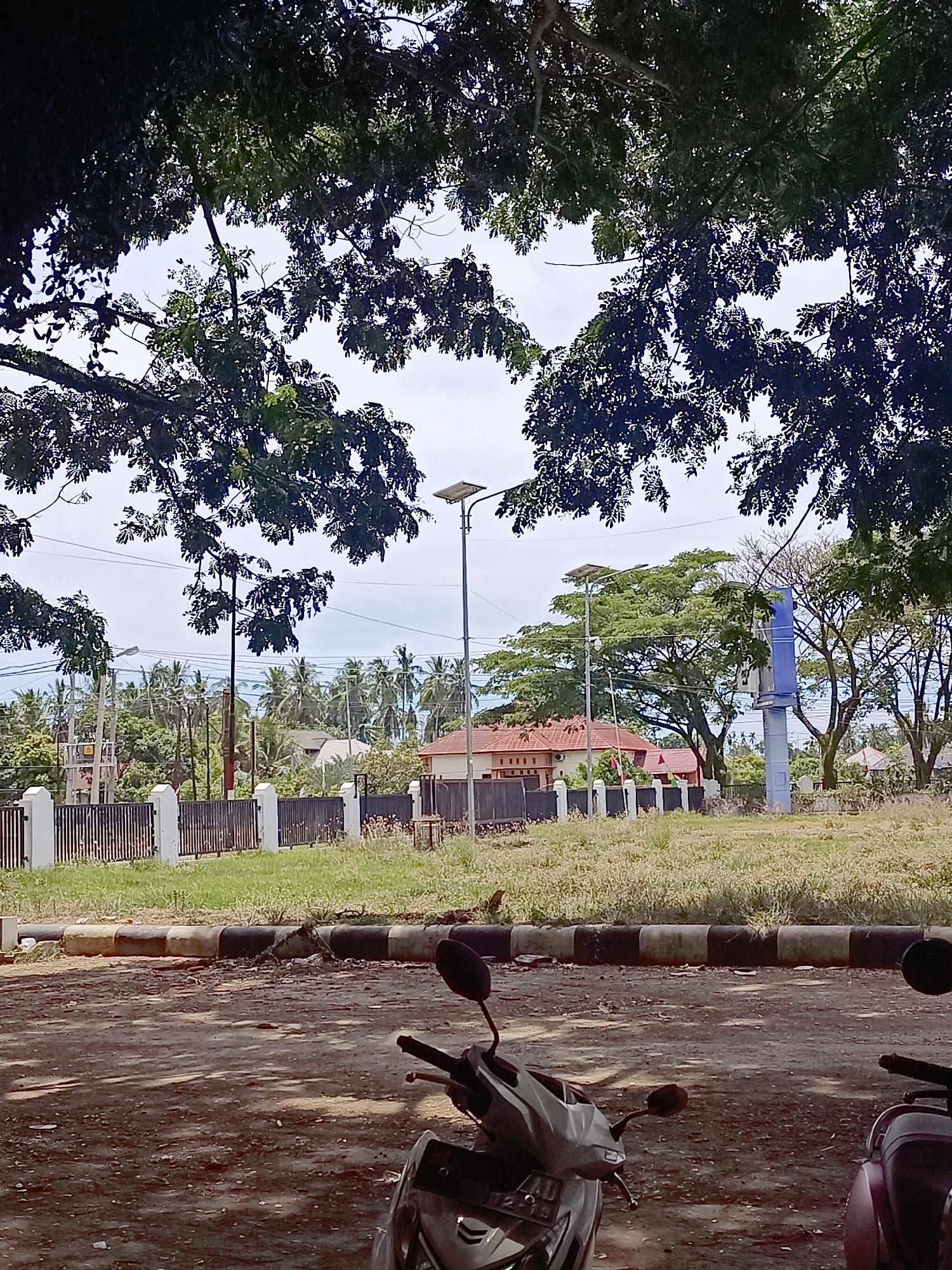 The image depicts an outdoor scene with a focus on the foreground and middle ground, framed by lush green foliage from overhanging trees. In the immediate foreground, a portion of a gray scooter is visible, facing left. To the right, the rear section of a dark-colored scooter is also present. The ground in the foreground is a dusty, uneven surface. A distinct curb, bordered with black and white painted sections, separates the foreground from the middle ground.
The middle ground features a neatly maintained area with a low, dark metal fence supported by white pillars. Beyond the fence, a grassy lawn extends towards a row of buildings with reddish-brown tile roofs, suggesting a residential or institutional area. A tall, blue pole with some signage is visible to the right of the buildings. In the background, a dense arrangement of palm trees indicates a tropical or subtropical environment. Power lines and streetlights are also present in the background, suggesting an urban or semi-urban setting. The sky is overcast with scattered clouds. The location is Meunasah Pupu, Indonesia.