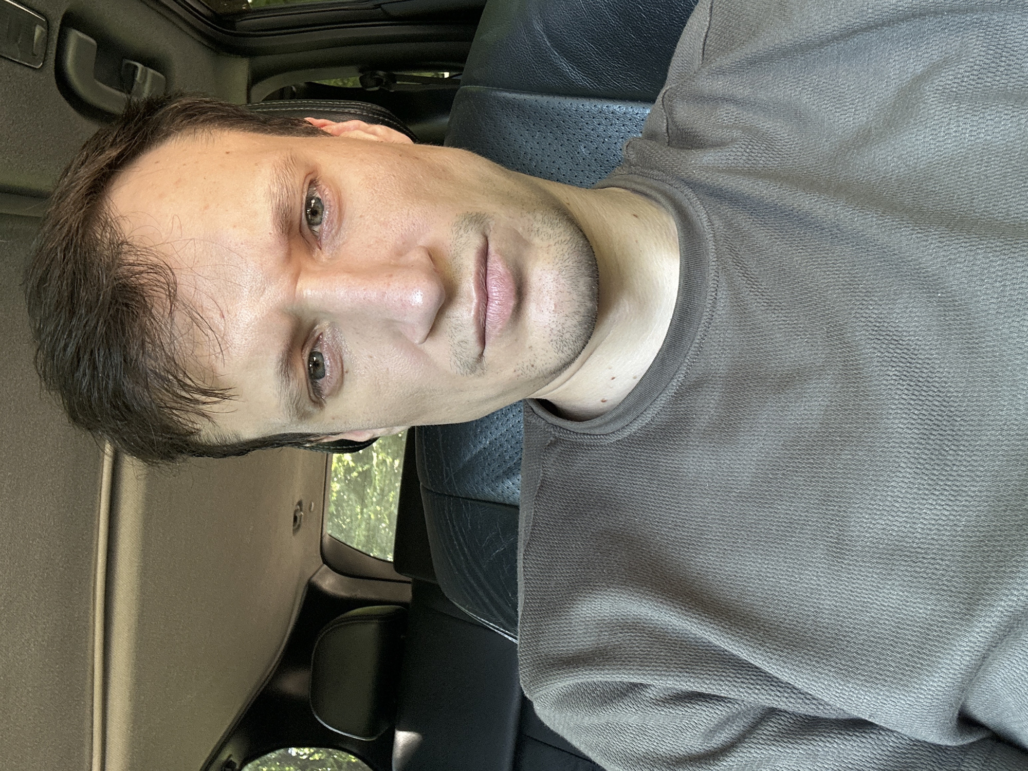 A man is sitting in the driver's seat of a car, looking directly at the camera. He has short, dark brown hair, fair skin, and green eyes. He is wearing a dark brown long-sleeved t-shirt. The interior of the car appears to be leather, with black seats visible behind him. Sunlight streams in from the windows, illuminating the scene. The background outside the car appears to be a wooded area, suggesting the car is parked in a natural or rural setting. There are no indications in the image that place this scene in Shatava, Ukraine.