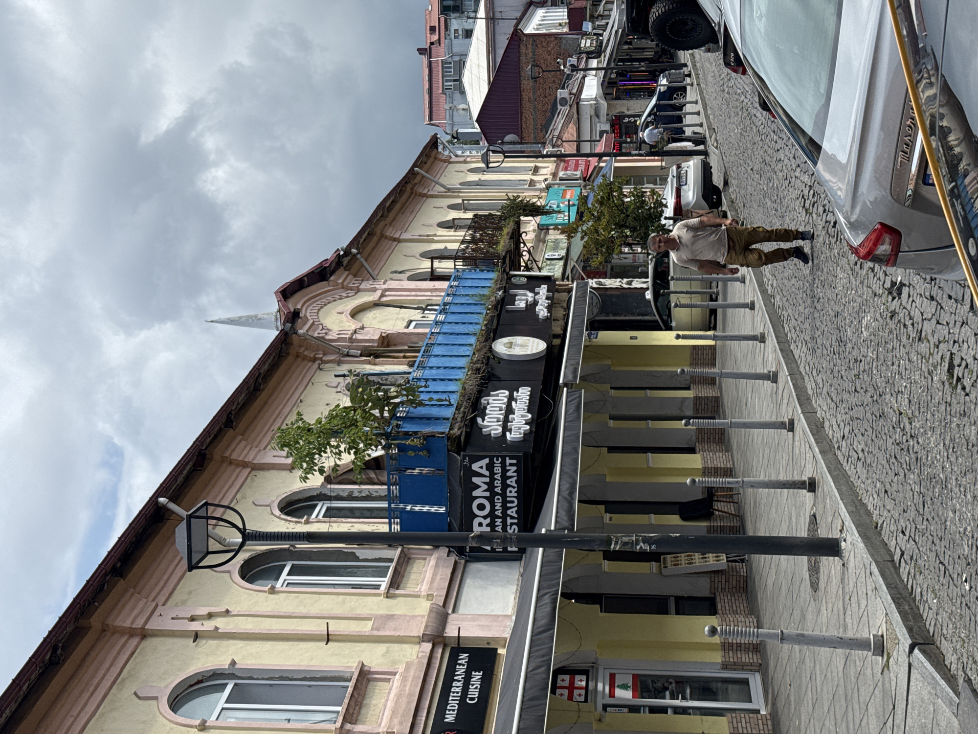 A man walks down a cobblestone street lined with buildings in Batumi, Georgia. The buildings have light yellow facades with pink trim and arched windows. Several businesses with awnings are visible, including one with a sign reading "MEDITERRANEAN CUISINE" and another with "ROMA IRAN AND ARABIC RESTAURANT". The man is wearing a light brown polo shirt and khaki pants. Cars are parked along the street, and a lamppost stands on the sidewalk. The sky is cloudy.