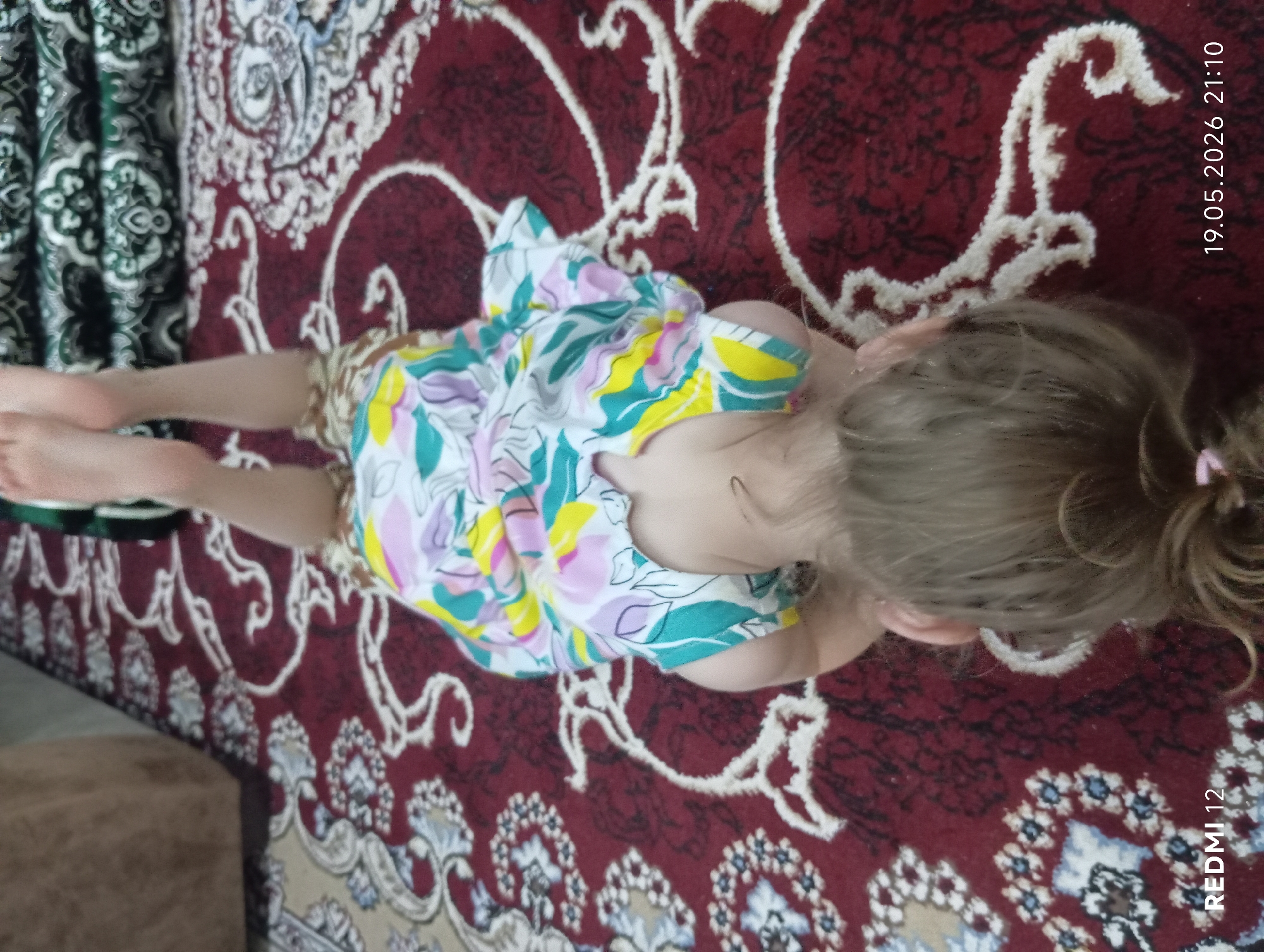 A young girl is lying on her stomach on a rug with a red and white pattern. She is wearing a floral print dress. Her hair is blonde and tied in a ponytail. The date "19.05.2026 21:10" is visible in the upper right corner of the image, and "REDMI 12" is in the bottom right corner. Part of a curtain can be seen on the left. Location is Bekabad, Uzbekistan.