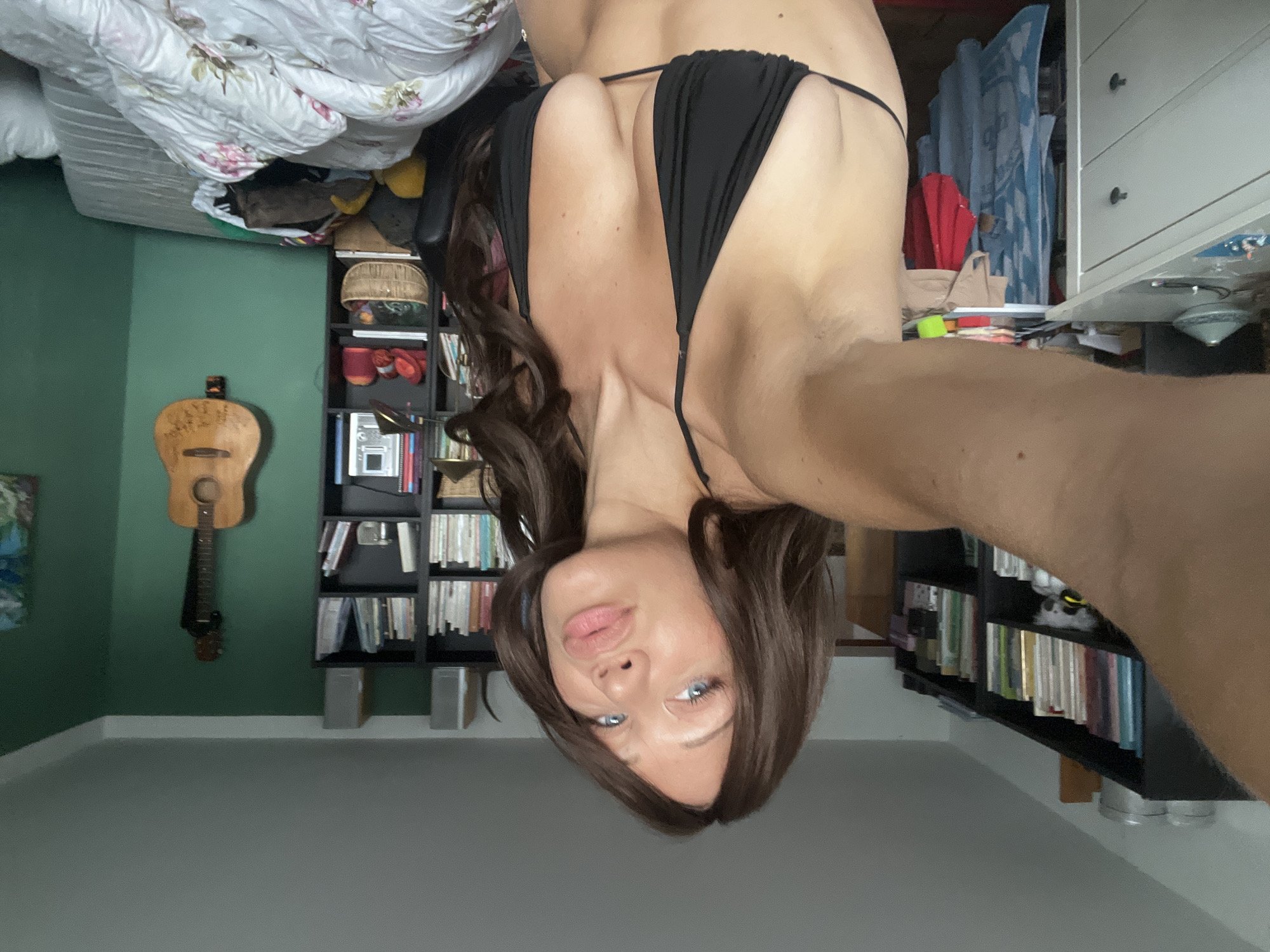 The media file depicts a person, likely a woman, in a bedroom. She is wearing a black bikini and her body is partially visible. The photo is taken from a low angle, with the subject's head near the bottom of the frame, implying she might be lying down or propped up in bed. The room appears to be cluttered, with a bed visible in the upper left corner, covered with white floral-patterned bedding. To the left, a dark green wall is adorned with an acoustic guitar. A black bookshelf filled with books is prominent on the right side of the frame, with another bookshelf visible behind the subject. A chest of drawers with light-colored drawers is also present to the right. The lighting suggests it is daytime, possibly late afternoon, with natural light coming from a window or overhead source. The overall atmosphere seems casual and personal, possibly a selfie taken in a private space. There is no visible text in the image.