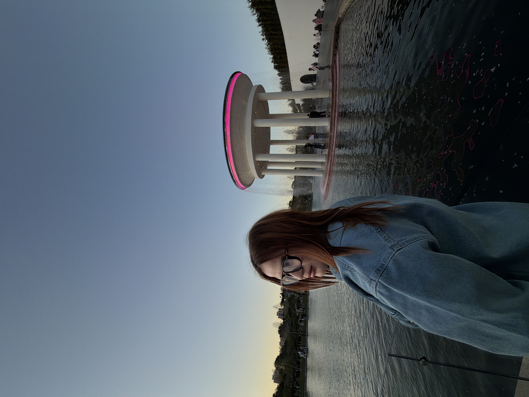 A young woman wearing glasses and a denim jacket stands near a body of water on a clear day. In the background is a modern architectural structure featuring a round, lighted roof supported by pillars, likely a fountain. The setting appears to be a park or public space. This is a location in Krasnodar, Russia.
