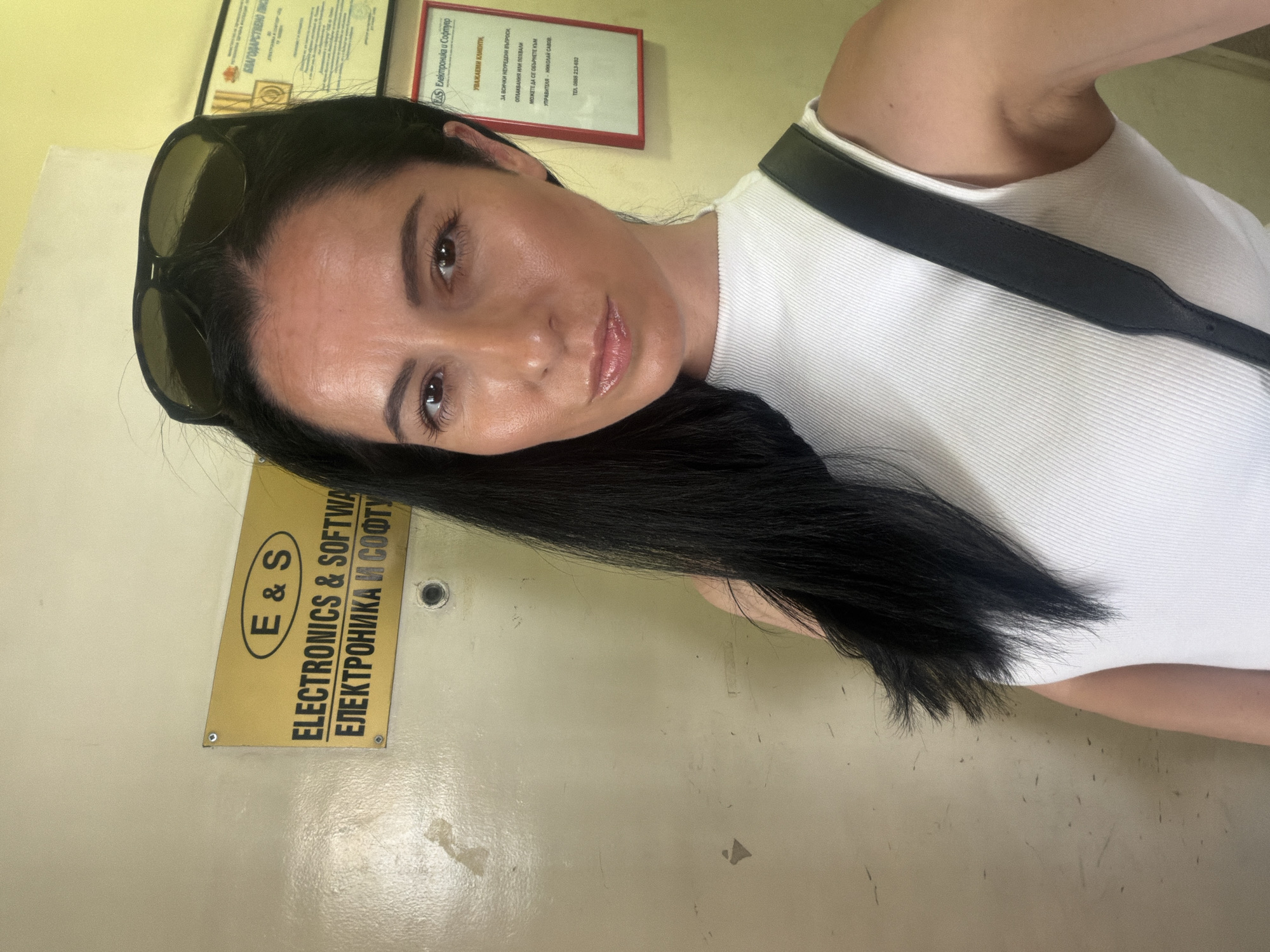 A selfie portrait shows a woman indoors, with dark hair and wearing a white top with a black strap. Sunglasses are perched on her head. Behind her are signs and documents affixed to the wall. One sign is yellow with black text: "E & S Electronics & Software, ЕЛЕКТРОНИКА И СОФТУ." Another sign reads in Bulgarian, "Електроника и Софтуер" indicating customer information including contact for "Nikolay Savov" in Pleven, Bulgaria.