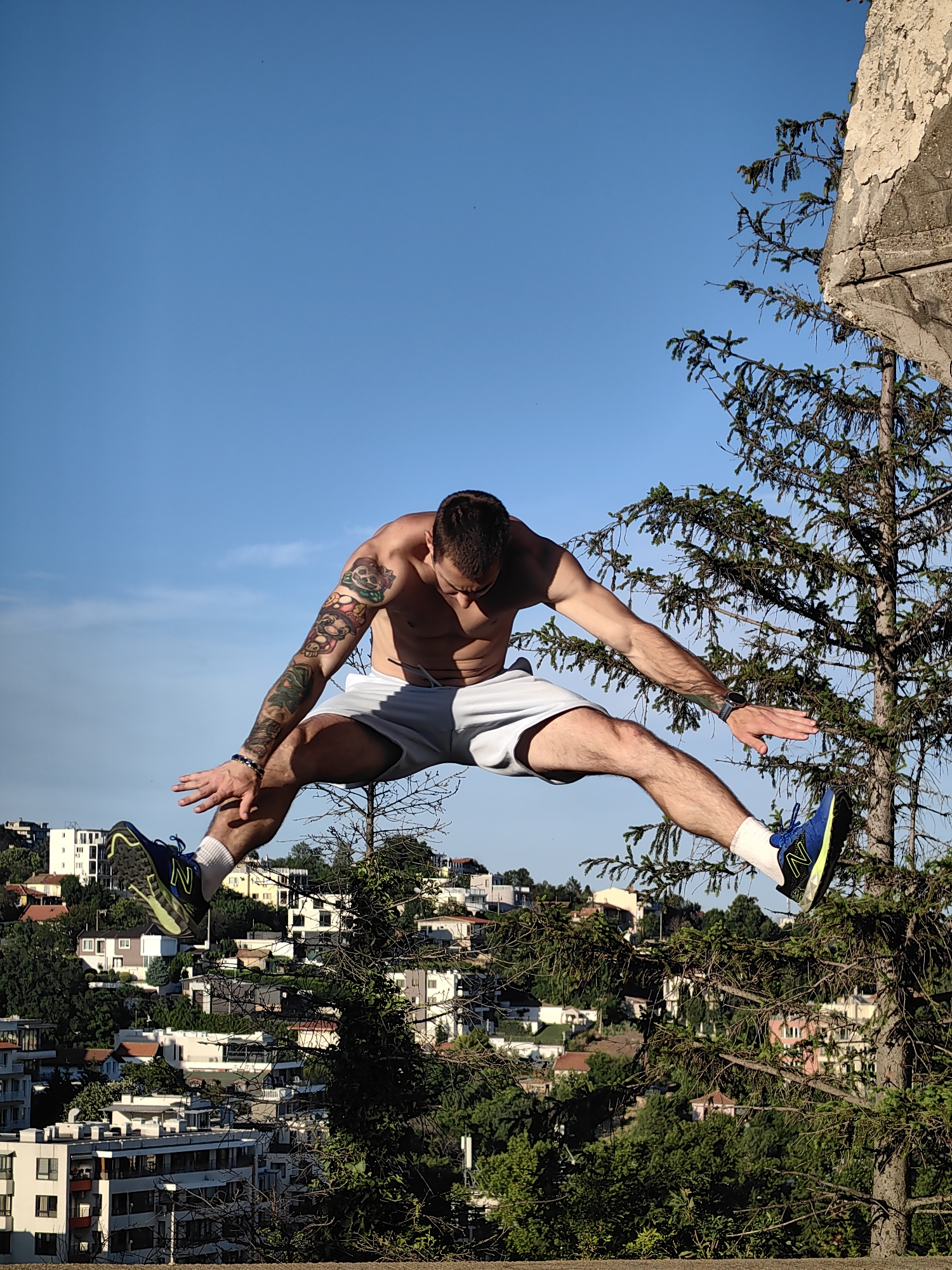A shirtless man with tattoos on his left arm is captured mid-air in a dynamic wide-legged jump. He is wearing light gray shorts and athletic shoes, one of which is clearly visible with a blue and neon yellow design. The man is positioned in the foreground, with a sprawling cityscape of Varna, Bulgaria, visible in the background. The city is nestled on a hillside, characterized by a mix of residential buildings with red-tiled roofs and modern apartments. Lush green trees and vegetation are interspersed throughout the urban landscape. A portion of a concrete structure, possibly a ruin or part of a building, juts into the frame from the upper right corner, alongside a dense evergreen tree. The sky is a clear, bright blue. The overall scene suggests an outdoor recreational activity or athletic training in a scenic urban environment overlooking the city.
