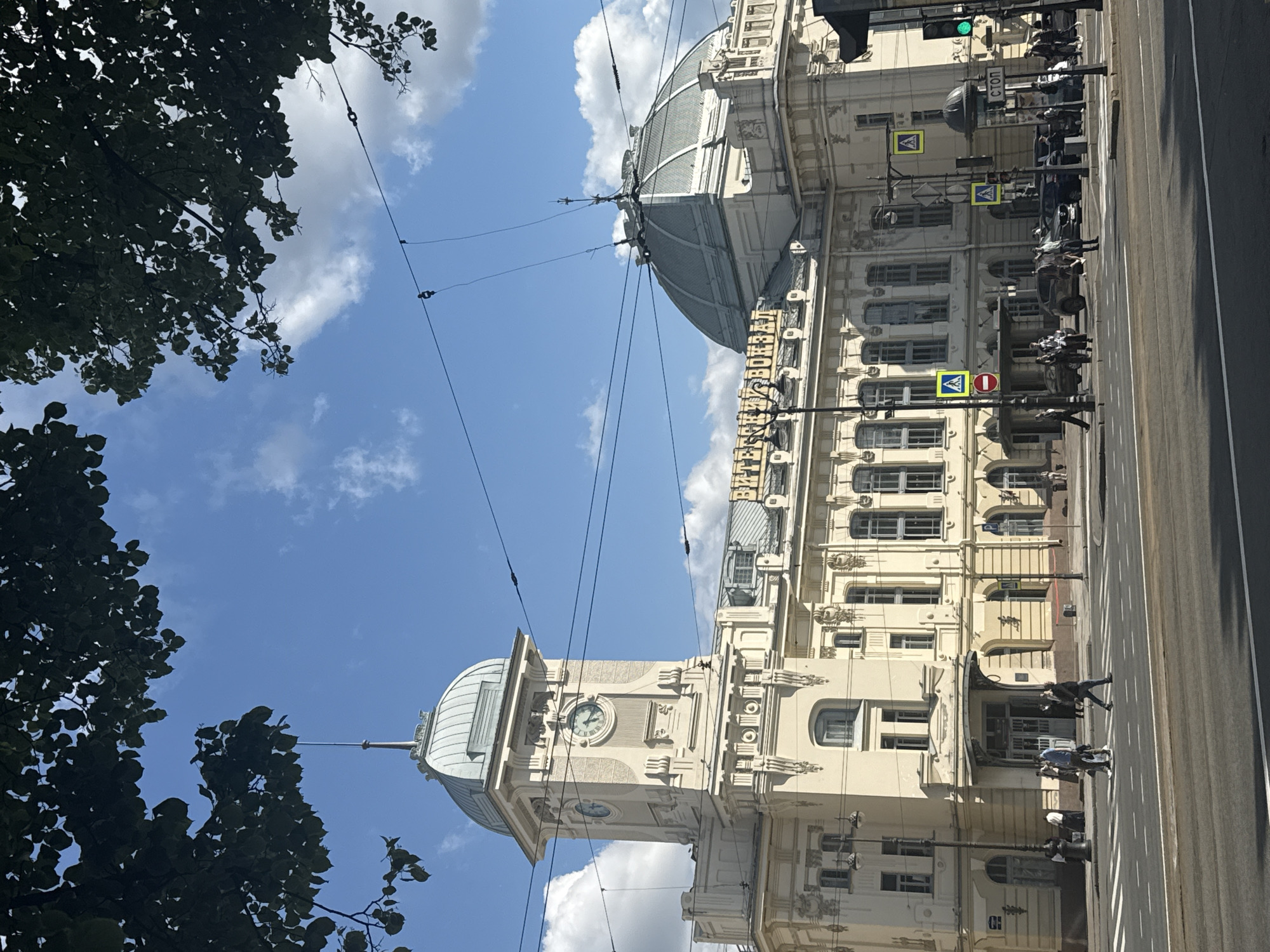 The image shows Vitebsky Railway Station, located in Saint Petersburg, Russia. The station is a grand, historic building with a prominent clock tower and a large domed roof. In the foreground, a street with a crosswalk is visible, with several pedestrians crossing and vehicles waiting. The sky is blue with scattered white clouds, and trees with dark green leaves are framing the top of the image. The scene appears to be a busy urban environment on a sunny day.