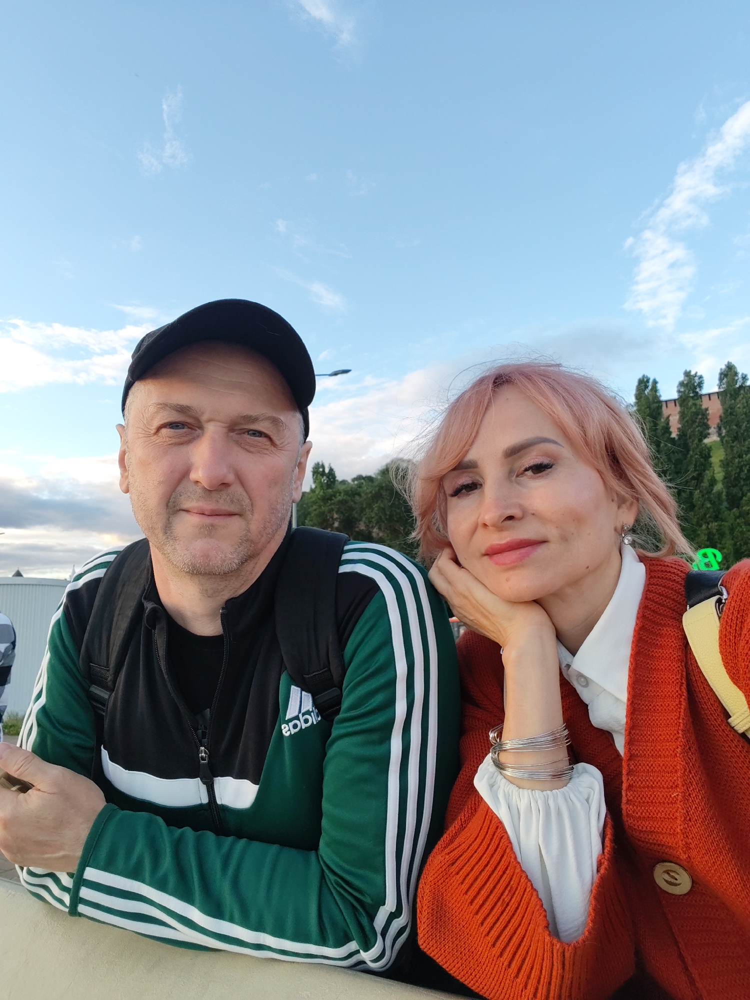 This is a selfie of a man and a woman outdoors. The man is in the foreground on the left, wearing a black baseball cap, a black t-shirt, and a green and black jacket with white stripes on the sleeves. He has a backpack on. The woman is to his right, with her head resting on her hand. She has light pink hair and is wearing a white shirt and an orange cardigan. She has several silver bracelets on her wrist.
The background shows a bright blue sky with some white clouds. To the right of the couple, there are tall trees and what appears to be a brick wall or building. The lighting suggests it is daytime, likely late afternoon or early evening due to the soft light. The overall mood seems relaxed and casual, with both subjects smiling faintly.
There is visible text on the man's jacket, which appears to be the Adidas logo. The location context is Nizhny Novgorod, Russia.
