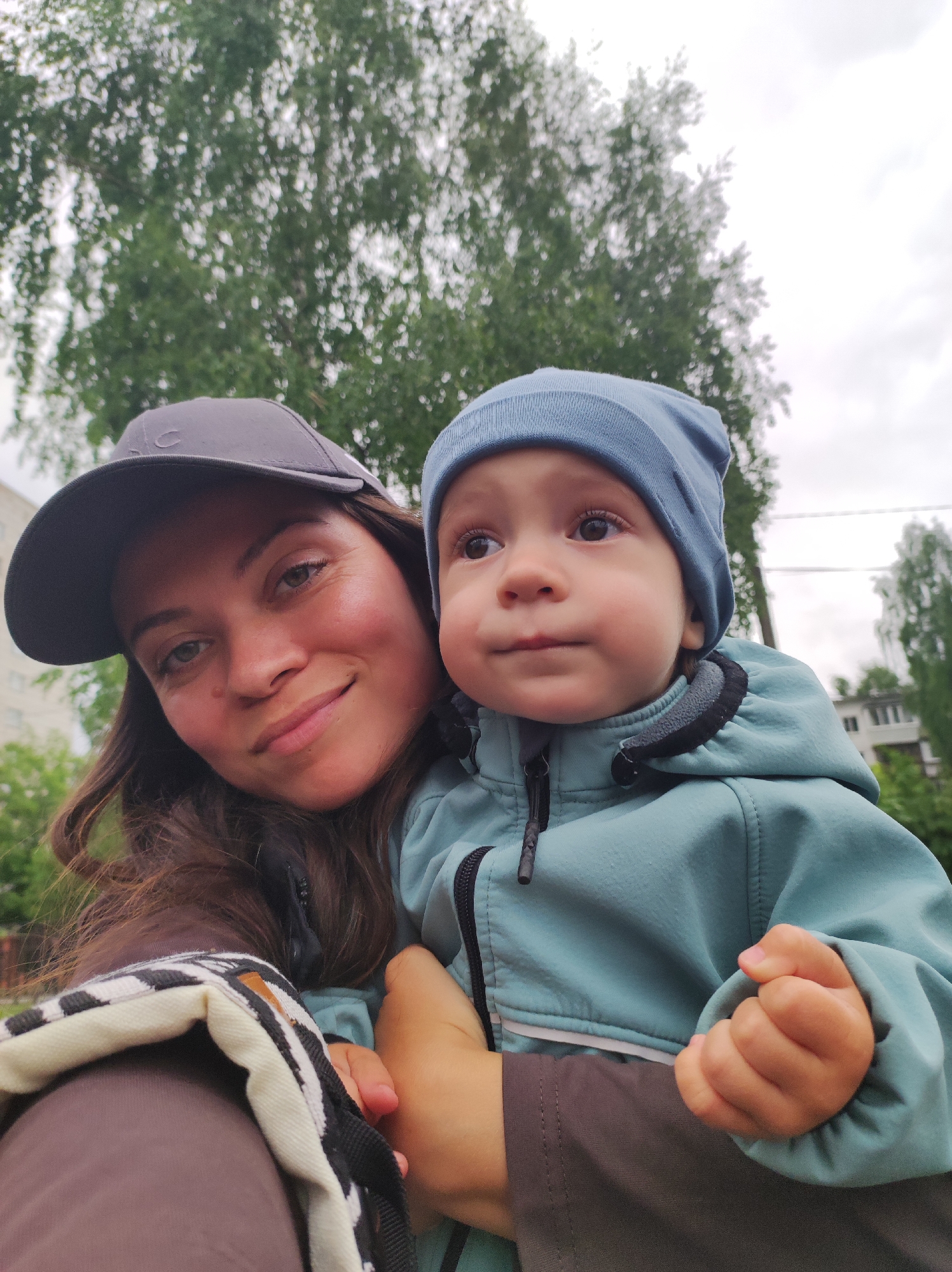 The image is a close-up, selfie-style shot featuring a woman holding a young child. The woman is wearing a dark baseball cap and has long, dark hair. She is smiling softly at the camera. The child is wearing a light blue beanie and a matching jacket. The child is looking off to the side, slightly away from the camera. The woman is holding the child close to her. The background consists of green trees and a cloudy sky. In the distance, a building is visible.
Based on the available information, it is impossible to verify the location of Chaikovsky, Russia in the scene, since the scene can happen elsewhere.