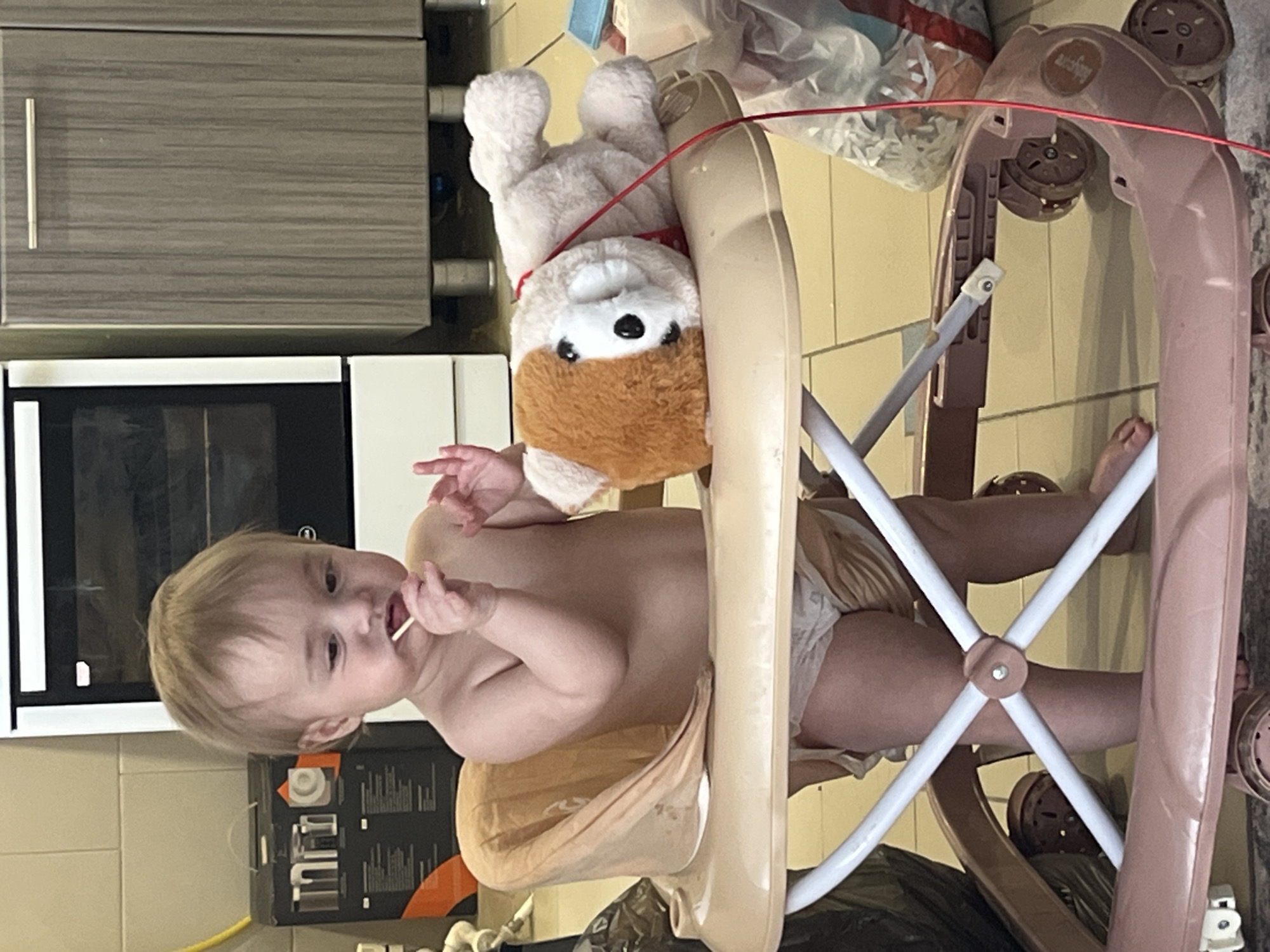 A baby is in a baby walker, holding a lollipop. A stuffed animal is attached to the walker. The setting appears to be indoors, possibly a kitchen, as cabinets and an oven are visible in the background. The exact location cannot be determined to be in Chaikovsky, Russia, without further information.
