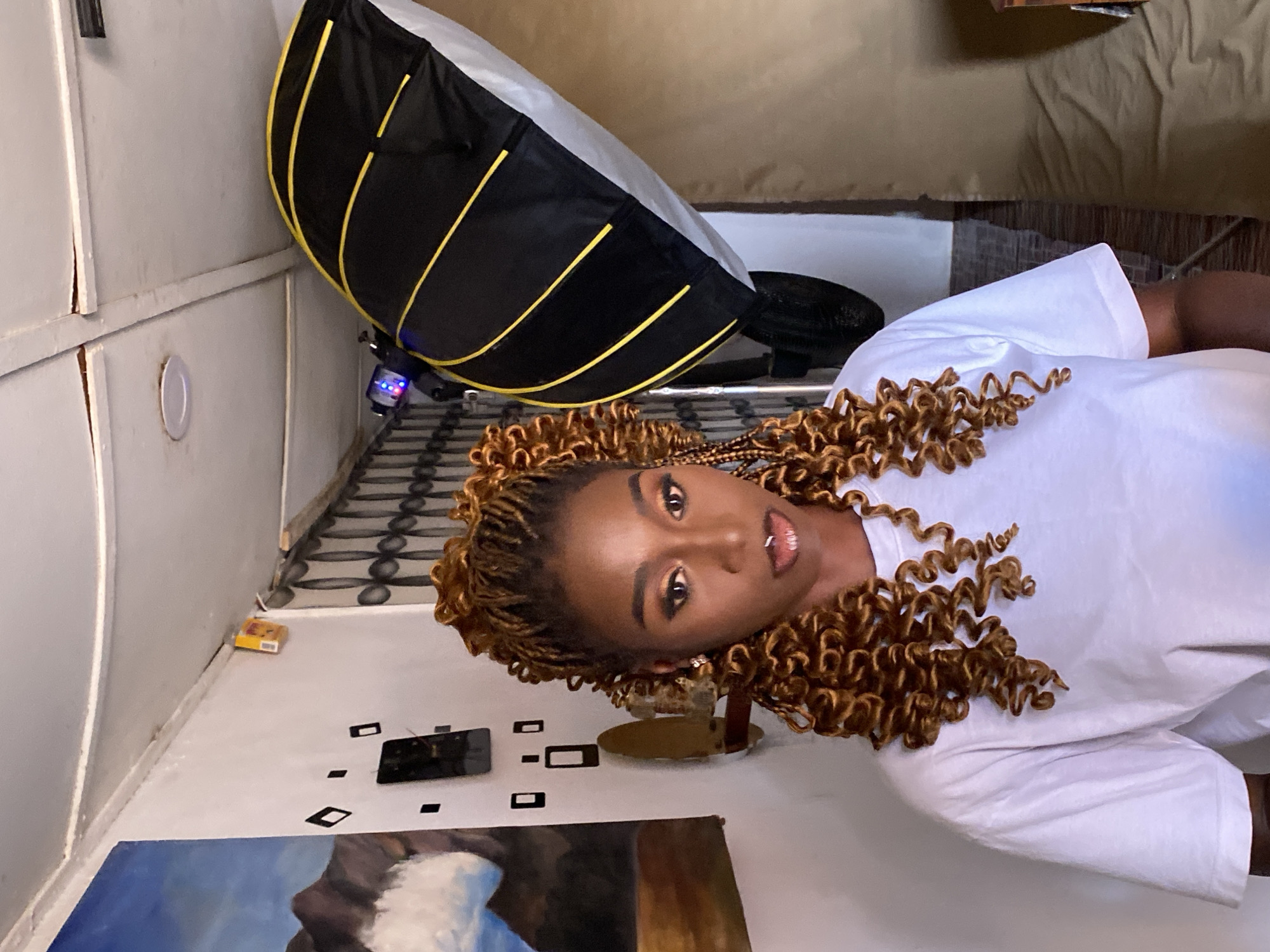 The image displays a young Black woman posing for a portrait in an indoor setting, likely a home or makeshift studio in Vom, Nigeria. The woman is positioned to look directly at the viewer, with her head slightly tilted. She has dark skin and her hair is styled in long, voluminous brown and blonde curly braids that cascade around her. She wears subtle eye makeup, a glossy lip, a nose piercing, and a labret piercing on her lower lip, and is dressed in a plain white t-shirt.
Behind and slightly to her left, a large black photographic softbox with distinctive yellow support rods is visible, mounted on a light stand with a small light fixture displaying blue and red LEDs. The background consists of light-colored walls, some showing cracks and paneling. The floor beneath her is light-colored, adorned with several black rectangular and diamond-shaped stickers or cutouts. A painting, featuring a blue sky and white clouds over a dark landscape, rests on the floor in the foreground, near a dark-colored smartphone. In a far corner, a small yellow box with illegible text is present. A patterned floor or rug with dark and light grey motifs extends into the background. Further back, a tan-colored piece of furniture, possibly a bed or couch, is partially visible. The scene suggests a casual photoshoot, with the woman as the central subject.