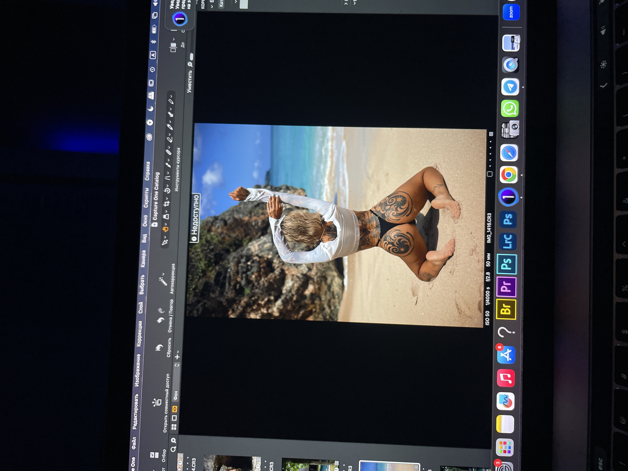 A photograph shows a laptop screen displaying a photo of a woman with tattoos on her back and thighs, kneeling on a sandy beach. The woman is wearing a white long-sleeved shirt and a black thong bikini bottom. She is facing away from the viewer, with her arms raised above her head. The background shows a tropical beach with turquoise water, a blue sky, and rocky formations. The laptop displays the editing software Capture One. The visible part of the operating system appears to be MacOS. There is no evidence indicating that the photograph or scene is related to the city of Kazan, Russia.