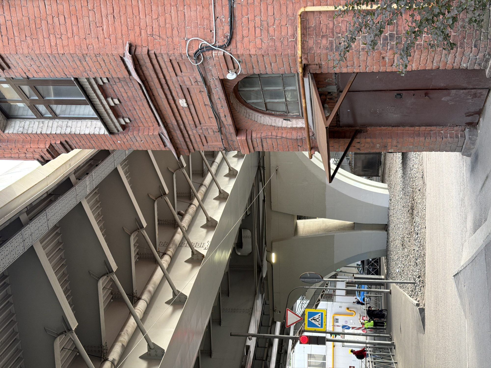 An urban scene depicts an underpass formed by a large, grey, industrial bridge or overpass, with a red brick building standing to its right. The bridge structure, occupying the left side of the image, features robust support beams, girders, and various pipes running along its underside, some with visible lights.
The brick building on the right has diverse fenestration, including a rectangular window with brown frames and a distinctive round-arched window with multiple panes. Exterior wiring and a small white security camera are mounted on its facade, alongside a yellow gas pipe running vertically. At its base, a dark metal door or gate, partially ajar, is nestled within the brickwork, partly obscured by green foliage from an overhanging tree.
At street level, beneath the bridge, there's a paved sidewalk and a strip of gravel leading to a road. A street pole holds a red traffic light, a triangular "Give Way" sign, and a blue square "Pedestrian Crossing" sign with a yellow border. Several people are visible: one individual in a red jacket near the traffic light, another in a yellow/green reflective vest pushing a cart, and other figures in the background. A blue car is also partially visible further down the road.
The scene captures a typical, mundane daytime moment in an urban environment, likely under overcast skies, with no specific event unfolding beyond daily pedestrian and street activity. No discernible text is visible in the image. This setting is located in Saint Petersburg, Russia.