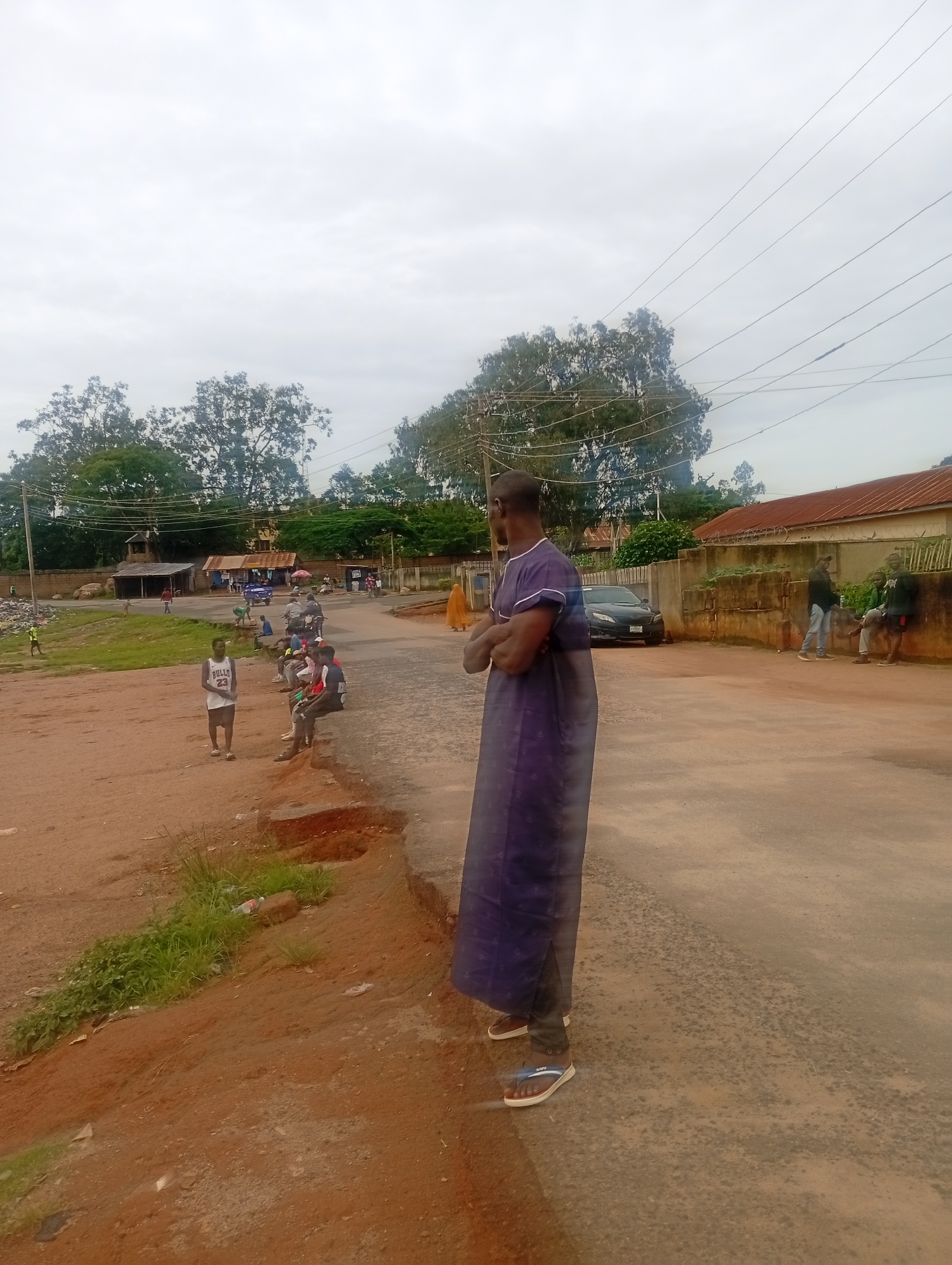 A medium shot captures a street scene in Jos, Nigeria, under an overcast sky. In the foreground, a man wearing a dark purple traditional outfit and white flip-flops stands facing away from the camera, arms crossed. To his left, a dirt path with some overgrown grass and a discarded bottle leads towards a group of people sitting on a raised curb. Among them, a young man in a white "Bulls 23" basketball jersey stands facing the camera. Further back, a few men are gathered near small roadside stalls, some on motorcycles. A black car is parked on the asphalt road. In the background, trees and buildings line the street, with electrical wires crisscrossing the sky. The overall impression is a snapshot of daily life in a semi-urban environment.