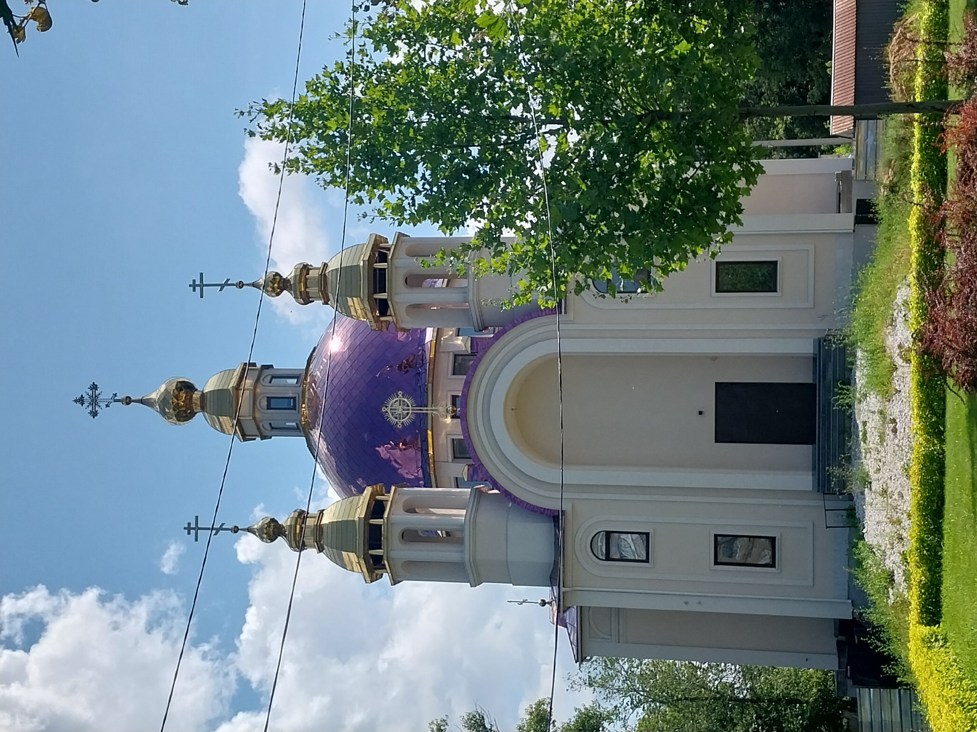 A white church with a large purple dome topped with a golden cross and smaller golden domes topped with crosses stands against a bright blue sky with scattered white clouds. The church has an arched entryway and several windows. A green tree with full foliage stands to the right of the church, partially obscuring it from view. In the foreground, there is a manicured lawn with bushes and a patch of red ornamental plants. Several power lines are visible stretching across the top of the image. The church is identified as the Church of the Intercession of the Mother of God, located in Dnipro, Ukraine.