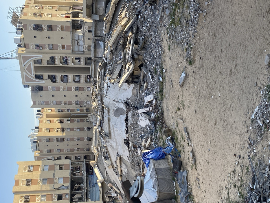 The image captures a scene of extensive urban destruction in Khan Yunis, Palestinian Territory, under clear daylight. In the foreground, sandy earth is scattered with various debris, including small rocks, concrete fragments, and twisted metal. To the immediate left, a light-colored canvas tent, appearing as a makeshift shelter, stands next to a collapsed blue tarp.
Dominating the midground is a massive, uneven mound of rubble, composed of broken concrete slabs, exposed reinforcement bars, and other structural debris, indicating the complete collapse of a building.
In the background, three multi-story residential buildings with light-colored, patterned facades are visible. These buildings exhibit widespread damage, including numerous shattered windows, visible holes and pockmarks on their exterior walls consistent with projectile impacts, and significant structural damage to balconies and lower floor sections, revealing destroyed interiors. Rooftops display multiple water tanks and communication equipment, such as satellite dishes and antennae. No individuals are present in the observable scene.