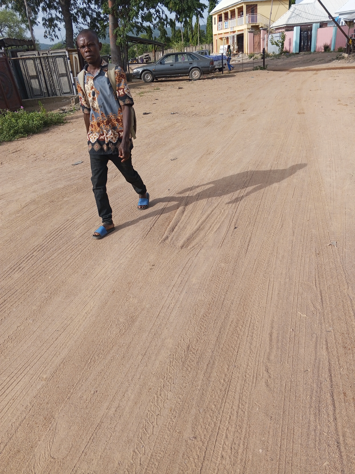 The image shows a man walking on a dirt road in Otokiti, Nigeria. The road is sandy with tire tracks. The man is wearing a patterned shirt, black pants, and blue sandals. He also carries a backpack. In the background, there are buildings, a parked car, and gates. The sky is overcast. Two other people are visible further down the road, near the car and buildings.