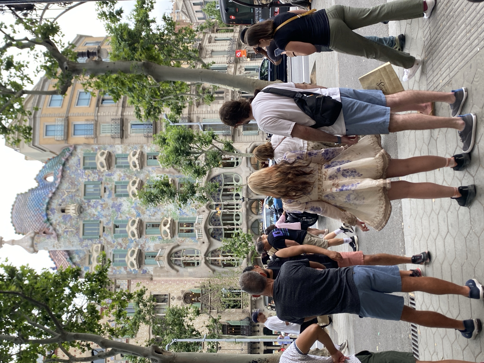 The image shows a group of people standing on a sidewalk in Barcelona, Spain, in front of Casa Batlló, a famous building designed by Antoni Gaudí. Casa Batlló is visible in the background with its unique architectural design. The people are facing towards the building. The street appears crowded. There are some trees along the street. A black bus is parked along the street.