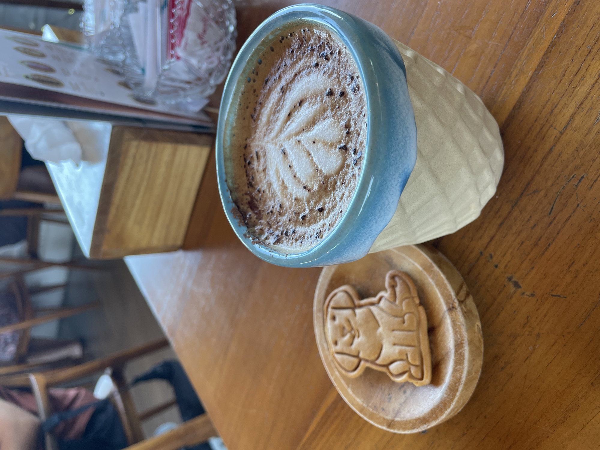 On a wooden table, a coffee cup with latte art sits on a small wooden coaster next to a cookie shaped like a character. A box of napkins and a container are also on the table. The location is Unknown, Unknown.