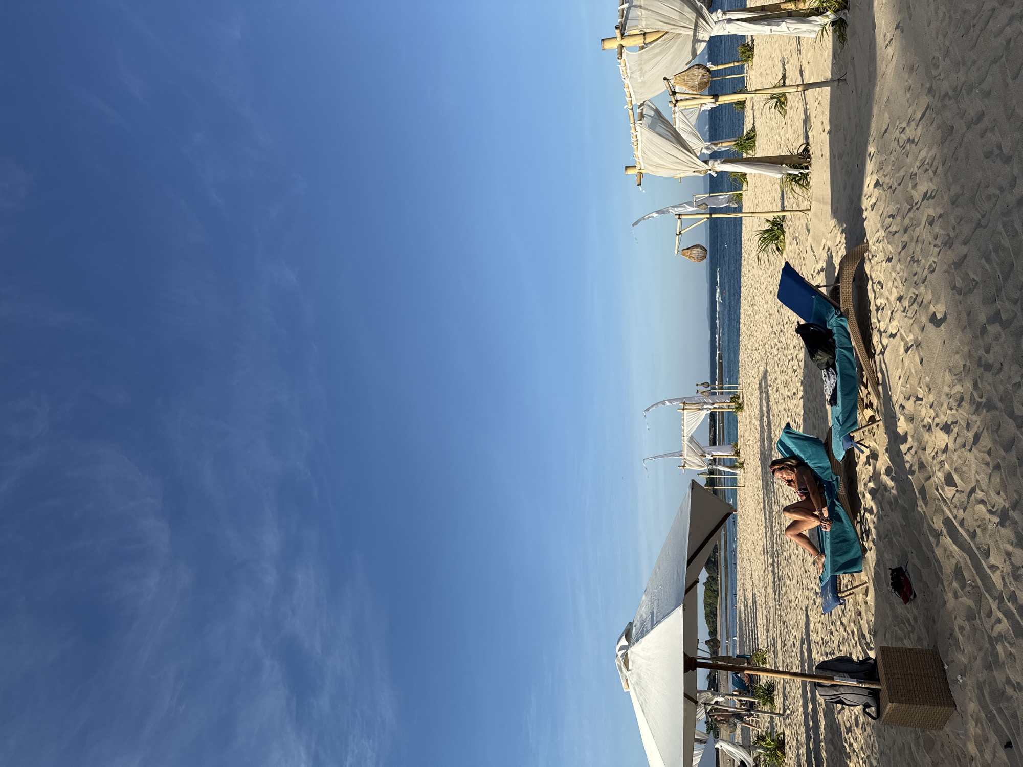 The scene is a sunny day on a beach in Kuta Selatan, Indonesia. A woman is reclining on a teal beach chair under a white sunshade. The beach is sandy and extends to the ocean, where a wave can be seen in the distance. A row of decorative white canopies stretches along the beach.