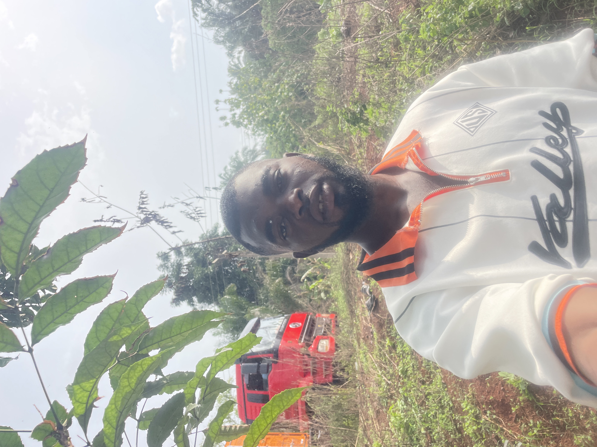 A man is standing in an overgrown field in Ogbe, Nigeria. He is wearing a white jacket with orange accents and the word "Valley" written in black script. Behind him, a red and yellow vehicle is visible amidst the vegetation. The background includes trees and a sky with high-hanging electrical wires. A leafy plant is in the foreground.