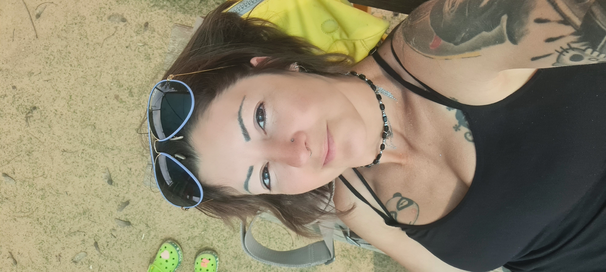 A woman is lying down in an outdoor setting. She is wearing a black tank top and has tattoos on her arm and chest. She has sunglasses resting on her forehead and a necklace around her neck. The ground appears to be sandy and there are green Crocs in the lower left corner of the frame. A yellow bag or cushion is behind her head. The photograph may have been taken in Lachish Regional Council, Israel.