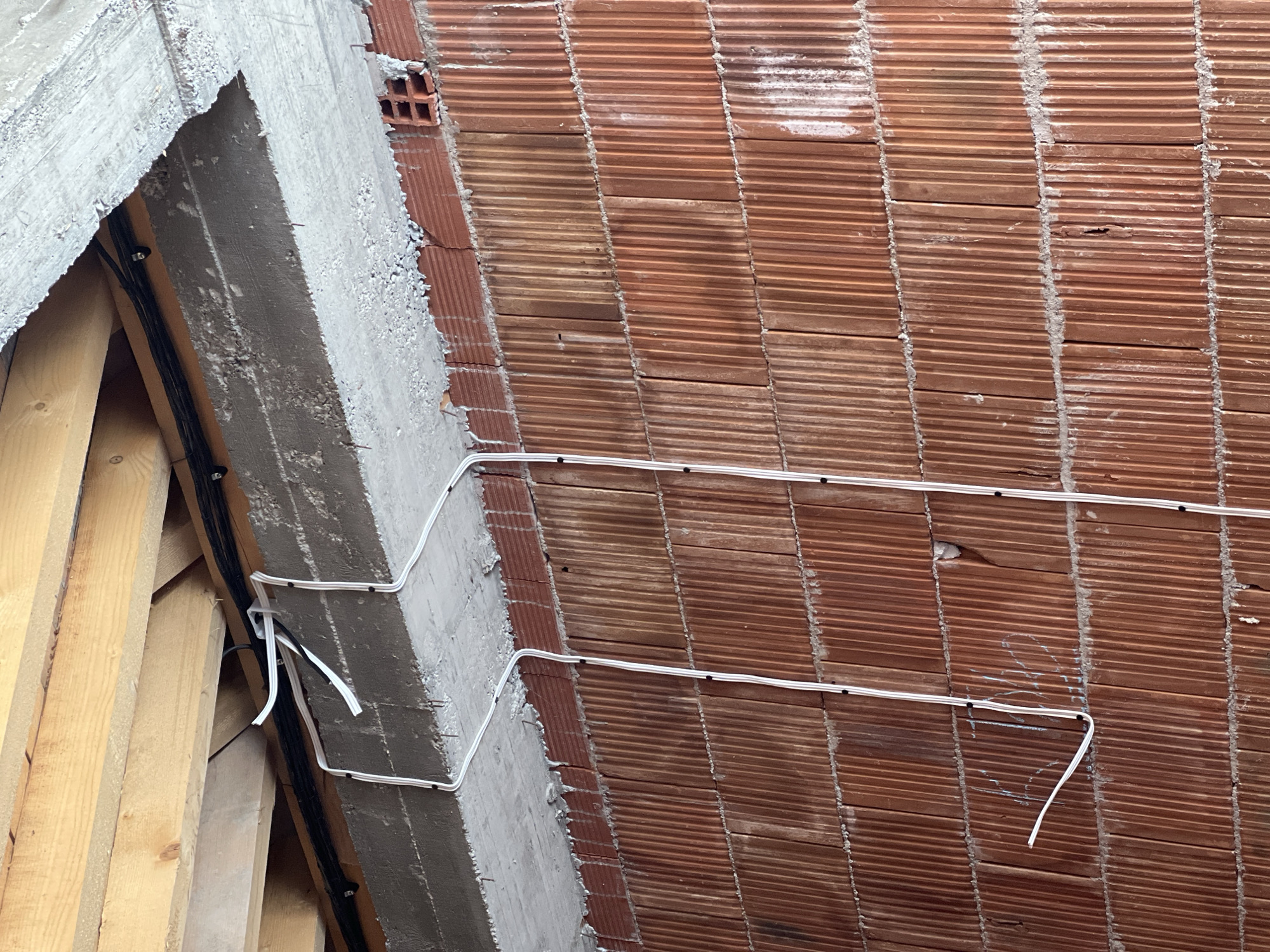 The image shows an unfinished ceiling construction with orange clay brick tiles arranged in rows, separated by cement. Two white electrical cables are affixed to the brick ceiling with black clips. To the left of the brick ceiling, a concrete beam runs along its edge, with black cables attached to its side near wooden supports. The scene is an interior view of a building under construction. While the location is specified as Topolovo, Bulgaria, there's no specific feature in the image that confirms this location definitively.