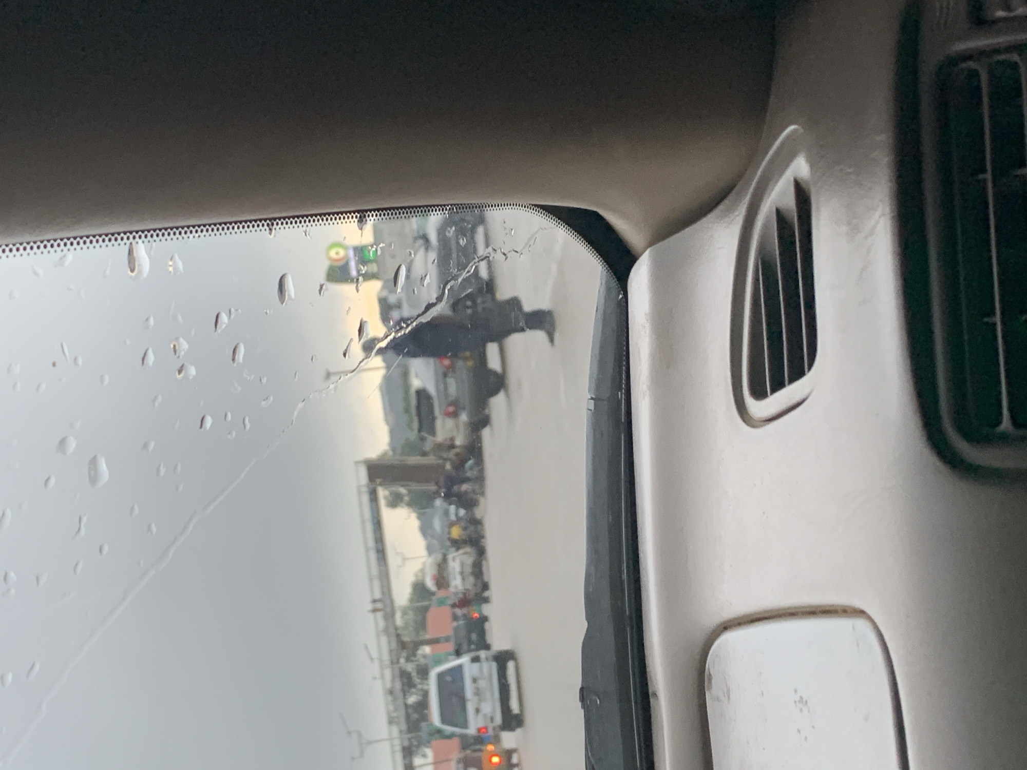 A man in a dark uniform and hat stands on a wet street in Gura Top, Nigeria. The view is from inside a vehicle looking through a rain-streaked windshield. The sky is overcast, and water droplets are visible on the glass, with a visible crack in the windshield. In the background, a pedestrian overpass is visible, along with several vehicles, including a white van and a darker SUV. A large green and orange billboard is also present in the distance. The interior of the vehicle shows a portion of the dashboard with an air vent and a storage compartment. The street appears to be a busy intersection with pedestrians and traffic.