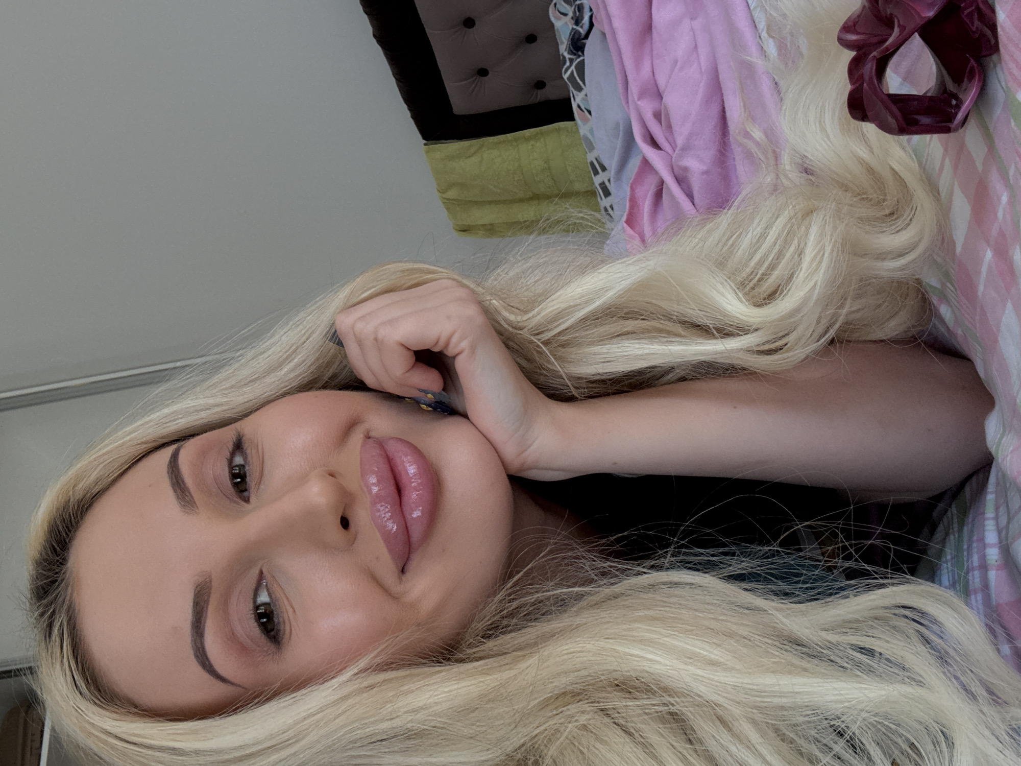 A close-up, selfie-style shot shows a blonde woman with long, wavy hair. She is lying on a bed, propped up on her left arm, which is resting on her chin. Her hair cascades around her face and over her shoulder. She is wearing makeup, including lipstick and eye shadow, and her nails appear to be painted. The background shows a bed with pink and white checkered sheets and a tufted headboard. A green towel hangs on the wall behind the headboard. A dark pink hair clip is visible on the bed in the lower right corner of the frame. The lighting suggests an indoor setting, possibly a bedroom.