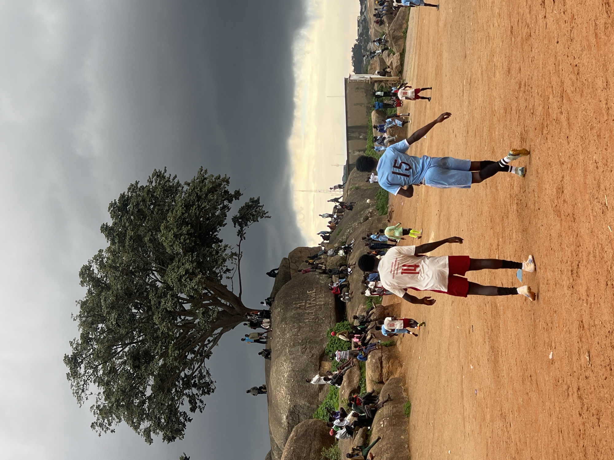 In Jos, Nigeria, a group of people are gathered on a rocky outcrop, with a large tree providing shade. Below them, on a dirt field, two young men are playing soccer. One player, wearing a light blue jersey with the number 15, is running towards the viewer's left, his right arm extended. The other player, in a white jersey with the number 14, is facing away from the viewer, also in motion. Several other figures, some in athletic wear, are visible on the field and on the rocks, suggesting a lively gathering or event. The sky is dramatic and overcast, with dark clouds creating a moody atmosphere. The terrain is predominantly orange dirt and large, weathered rocks, characteristic of parts of Jos.