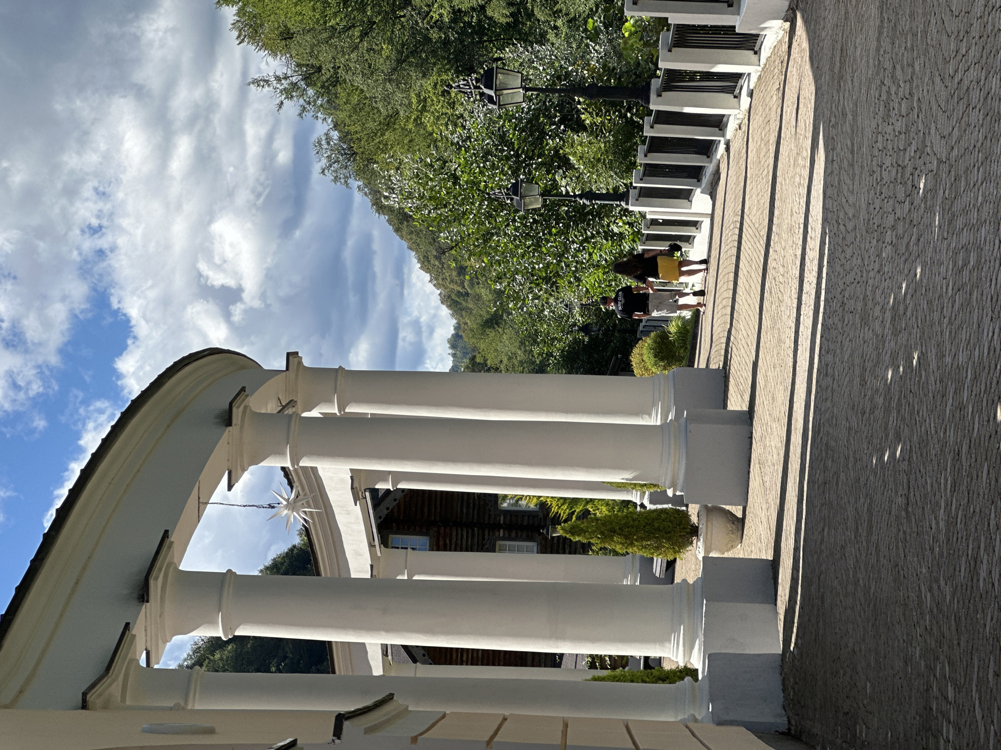 A couple walks away from the camera on a sun-dappled, cobblestone path bordered by a white fence with black metal railings and lampposts, in Estosadok, Russia. They are walking under a curved colonnade with white columns, leading towards a dense, green forest on a hillside under a cloudy sky. The woman is wearing a yellow skirt and the man is wearing shorts and a t-shirt. A white, star-shaped decoration hangs from a chain from the colonnade's ceiling.