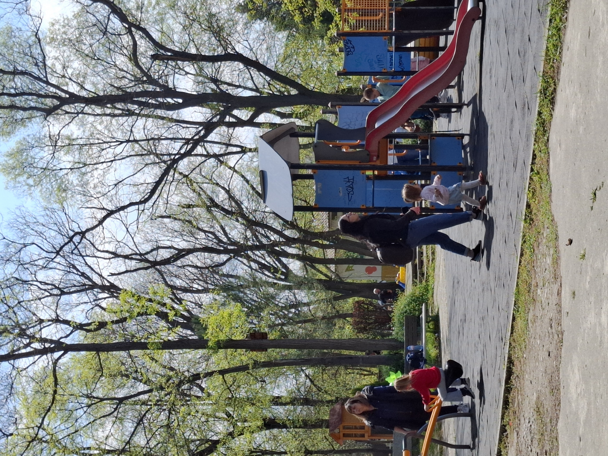 The image depicts a park scene in Plovdiv, Bulgaria. Dominating the foreground is a playground with a red slide, blue and orange structures, and a climbing frame. Several individuals are present, including adults and children, engaged in various activities. One child appears to be descending the slide, while others are near or on the playground equipment. A woman is present near the play structure, possibly supervising the children. Further back, other individuals are visible, sitting on benches amidst trees and greenery. The surrounding environment consists of trees, grass, paved walkways, and glimpses of buildings. The sky is visible through the trees.