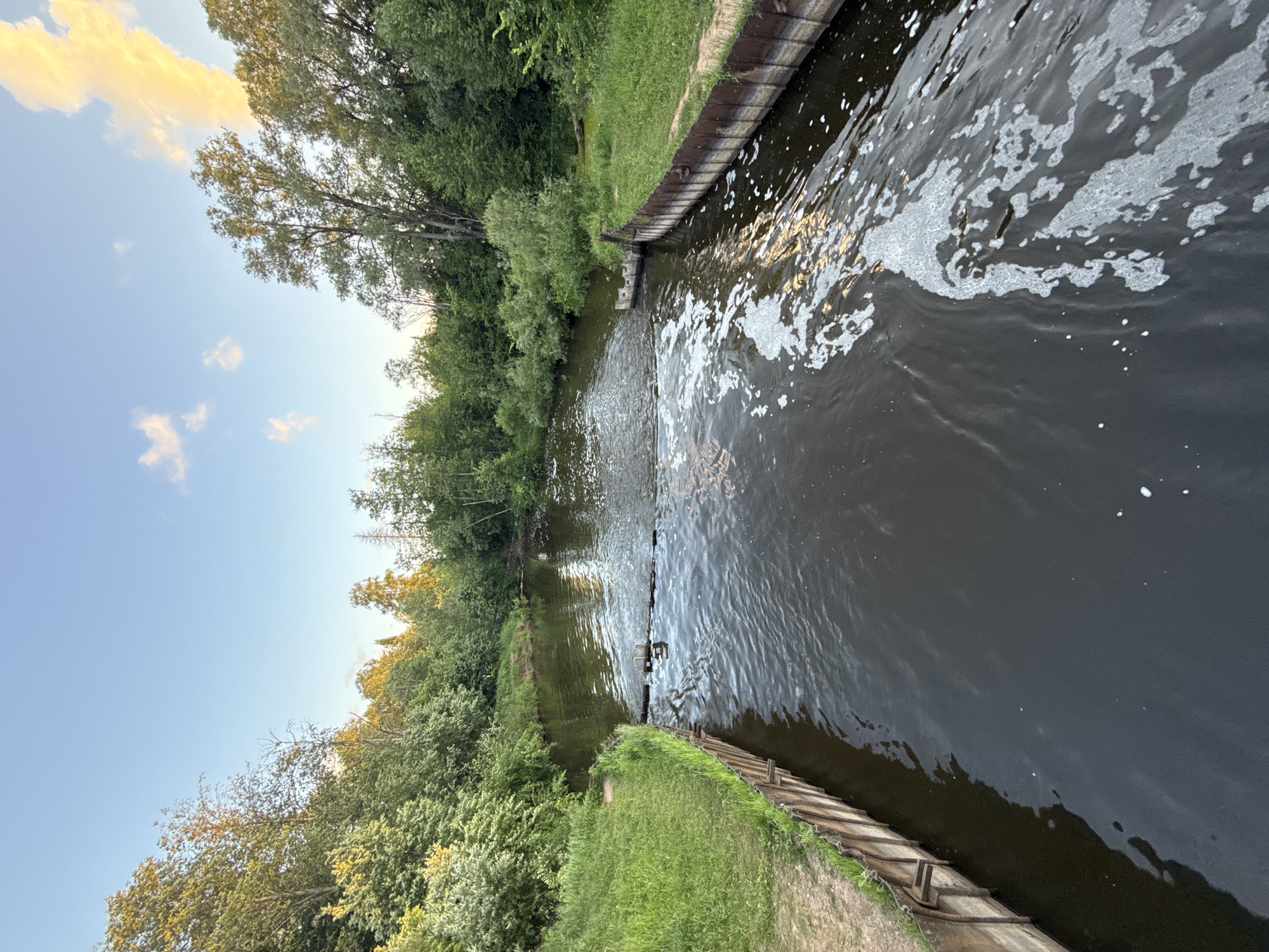 The image depicts a tranquil river scene with lush green vegetation lining its banks. The river flows calmly, with a section of wooden structures and a gentle cascade creating a textured surface on the water. In the foreground, foamy water churns, indicating movement or a subtle disturbance. The sky above is a soft blue, with scattered clouds tinged with the warm hues of the setting sun, suggesting it is late afternoon or early evening. The overall atmosphere is serene and natural, characteristic of a rural or parkland setting. This scene is located in the city of Rusalevka, Russia.