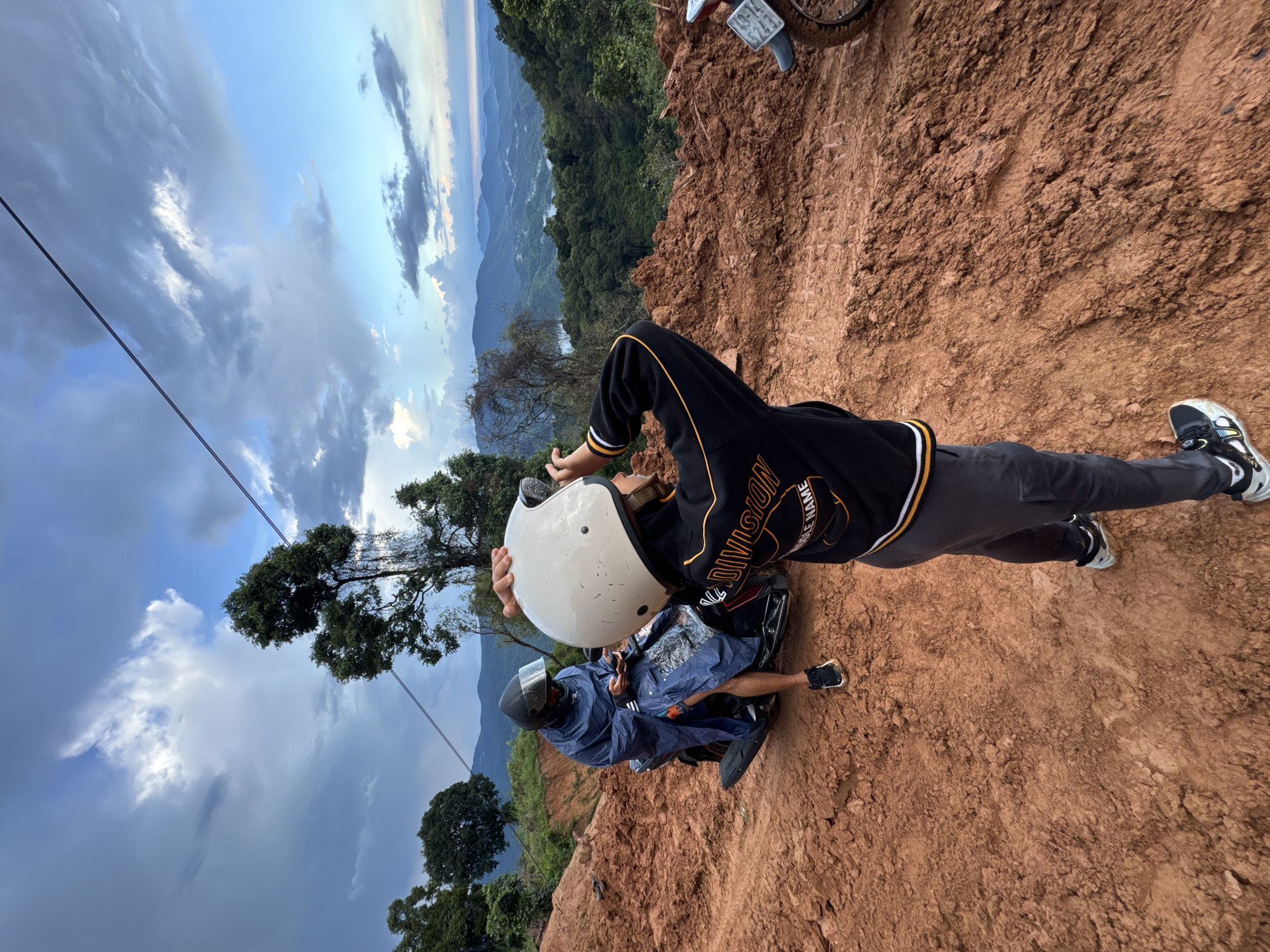 This is a medium shot, full-color, outdoor photograph taken from a low angle, looking up at two people and a scenic mountain landscape. The foreground is dominated by a muddy, reddish-brown dirt road, with tire tracks visible. To the left, a person wearing a blue rain poncho and a black helmet sits on a dark scooter. They are looking down at their phone. In the center and right foreground, another person wearing a black and yellow jacket and a white helmet stands with their back to the camera, holding the helmet up to their head as if to take a photo. The background features a lush, green mountain range with several layers of hills receding into the distance, under a sky with dramatic clouds and hints of blue. Power lines are visible stretching across the sky. The scene appears to be on a mountain road, possibly in a rural area. While the image does not explicitly depict Nao Sẻ, Vietnam, the landscape is consistent with mountainous regions in Southeast Asia.