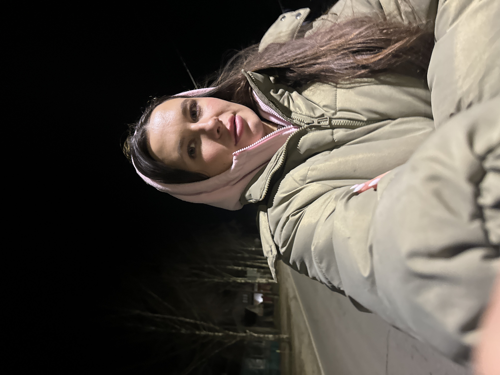 A person wearing a tan puffer jacket over a pink hooded sweatshirt is posing for a selfie outdoors at night. Trees line the background, appearing dark against the night sky. The location may be Красный Маяк, Russia.
