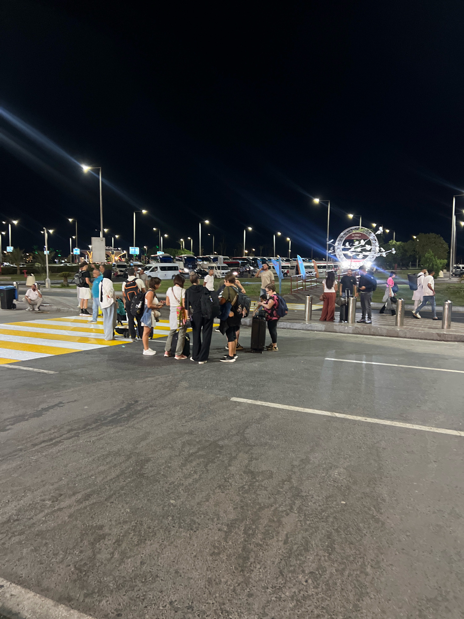 The image depicts an outdoor scene at night, likely at an airport or transportation hub in Sharm El Sheikh, Egypt. A group of people, appearing to be travelers, are gathered near a pedestrian crossing marked with yellow and white stripes. Many individuals are carrying luggage, suggesting they are arriving or departing. Various vehicles, including vans and buses, are visible in the background, illuminated by streetlights. A large, circular decorative structure with lights is also prominent in the middle ground. The overall environment appears to be a well-lit public space, with a dark sky overhead.