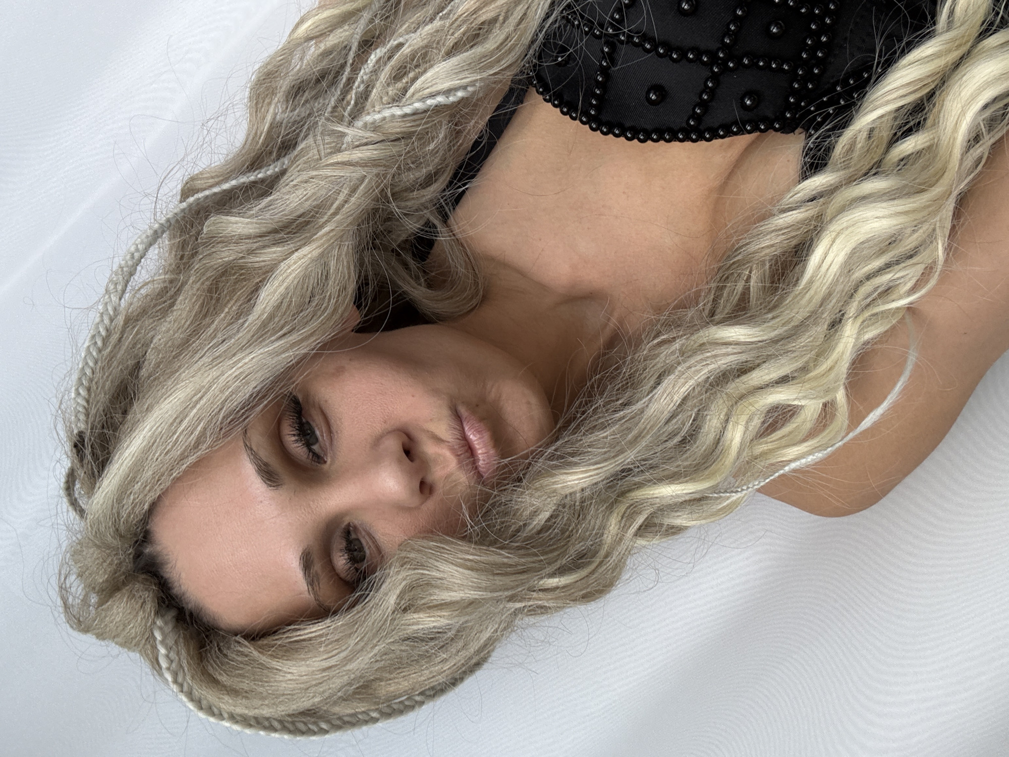 A close-up, portrait-style photograph shows a woman with long, blonde, wavy hair lying on a white surface. The woman's face is the focal point, with her eyes partially closed. She is wearing a black top with small, beaded embellishments near her neck. The background consists of a plain white material. The photograph does not provide any information about the location or city of Rusanovo, Russia.